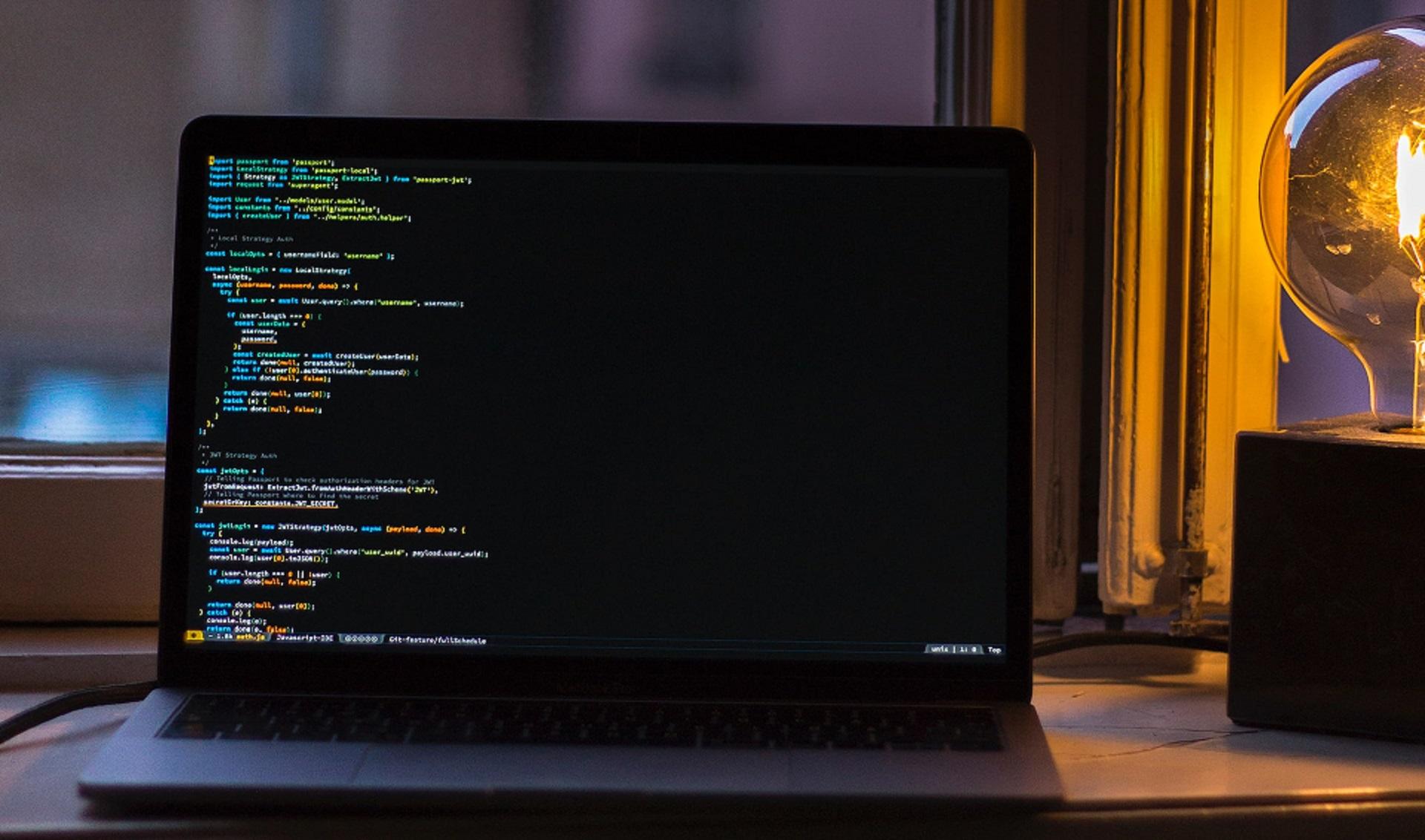 An open laptop computer displaying lines of code sitting next to a light casting a a warm amber glow.