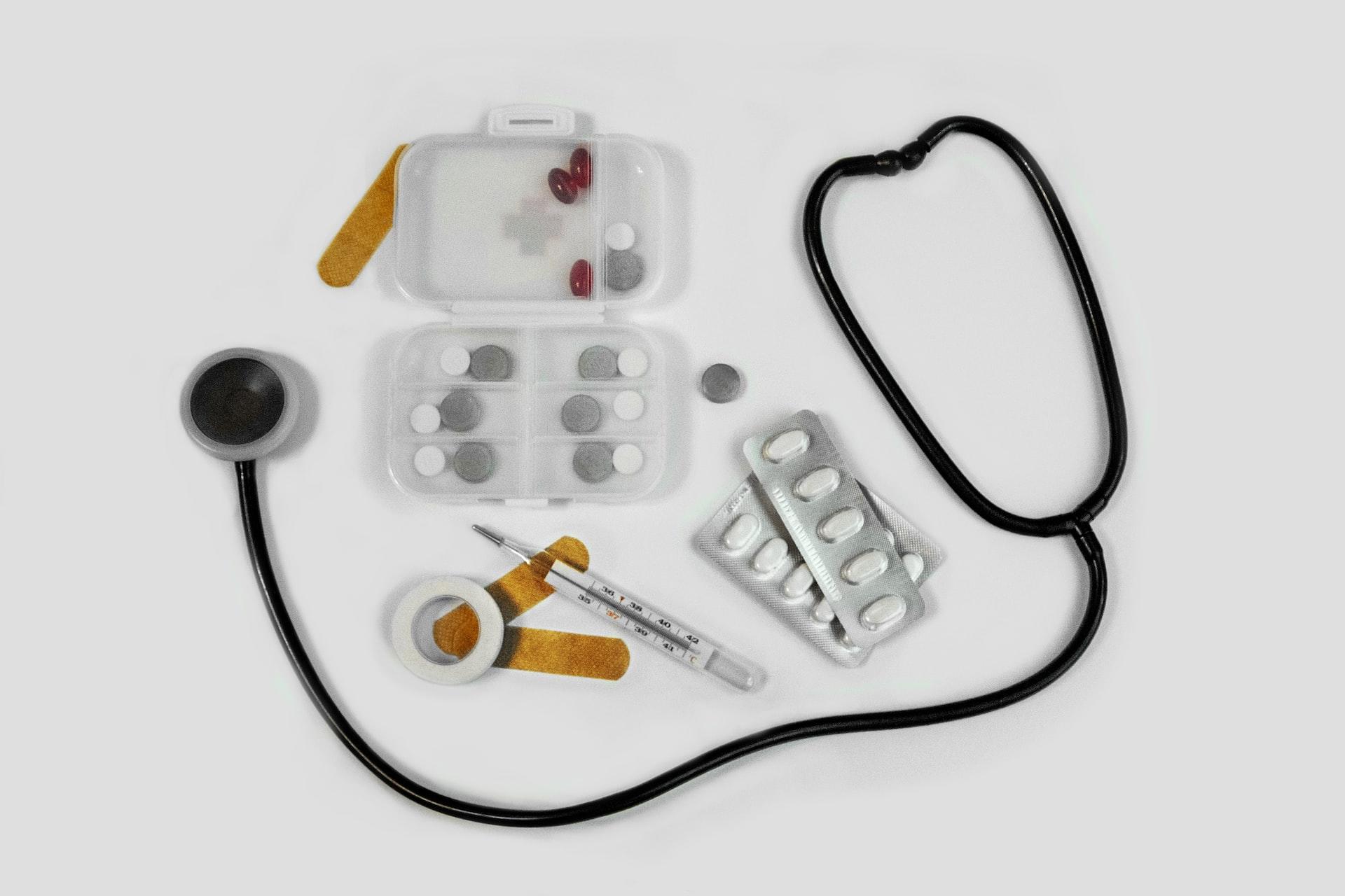 A stethoscope laying on a white surface, bracketing assorted medical staples like pills, a thermometer and other devices.