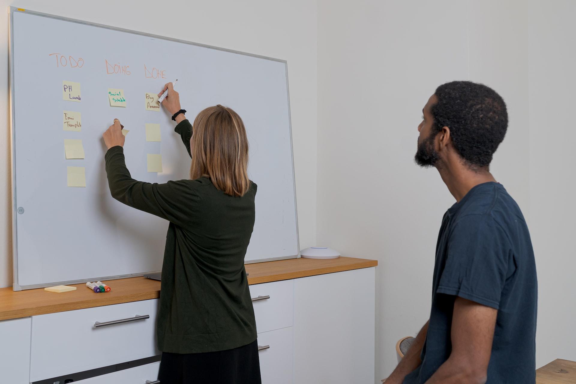 A woman in business attire affixes sticky notes to a whiteboard while a man in a dark-coloured shirt looks on.