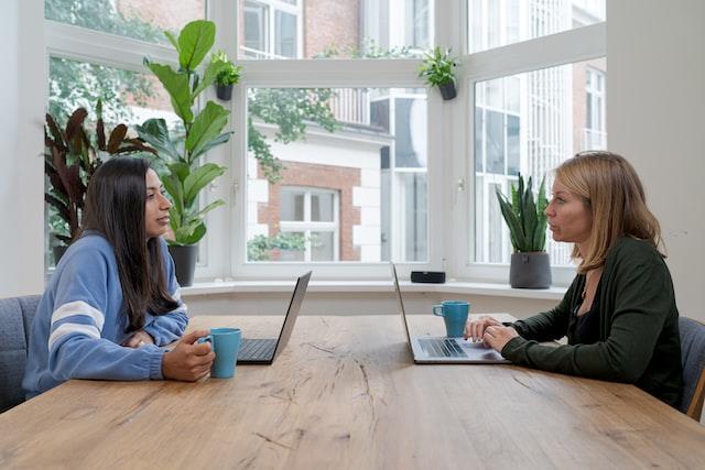 A woman in a dark-coloured top sits at a wooden table in front of an open laptop, talking to a woman in a light-blue top who is holding a blue coffee mug.