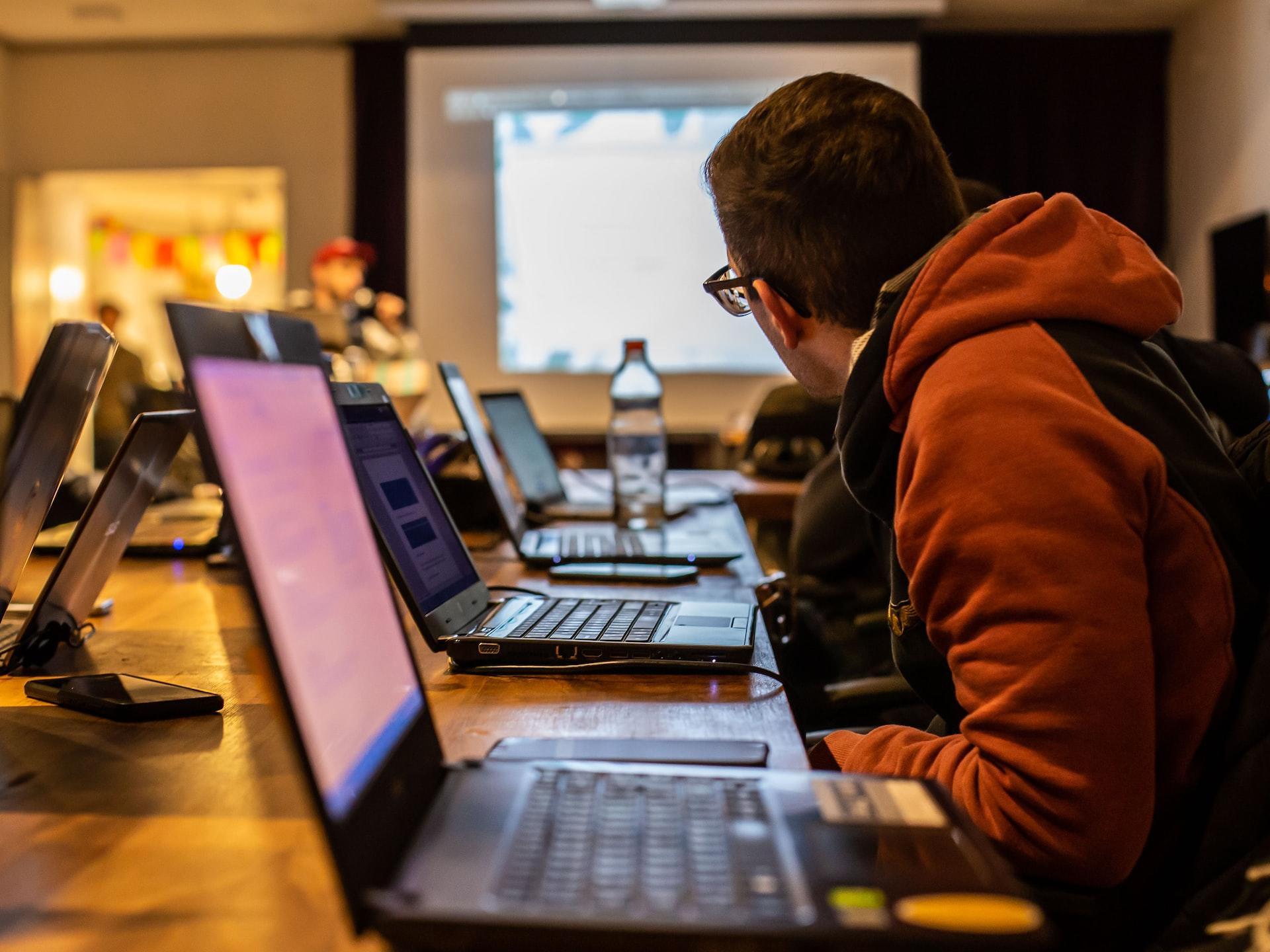 A student in a red jacket, with an open laptop in front of him, leans forward to see the classroom's display screen on a faraway wall. 