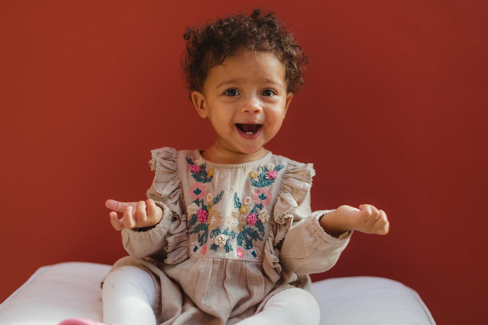 A toddler wearing a brightly coloured dress and white tights holds her hands, palms-up, while smiling. 