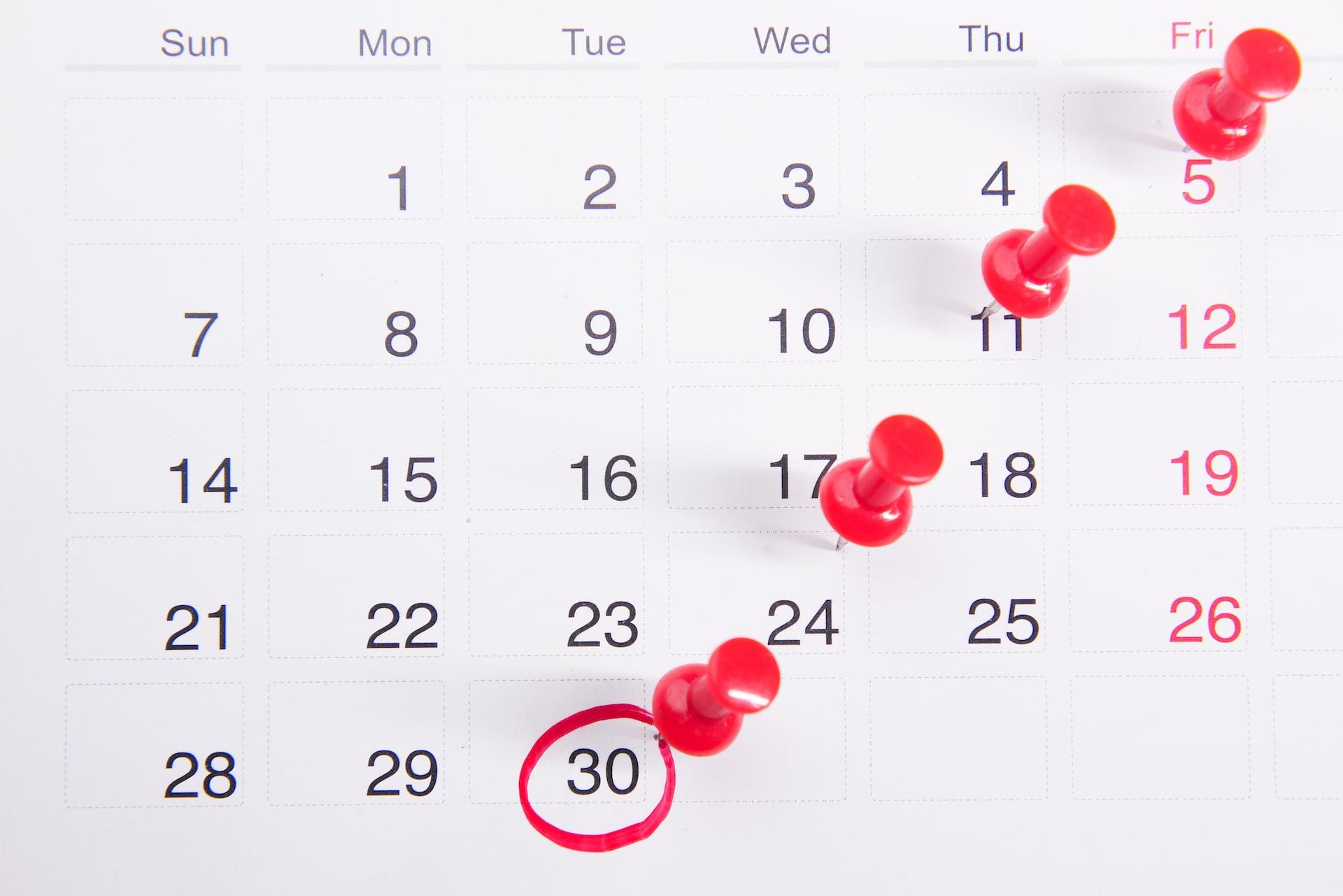 A white wall calendar page with the days printed in black ink with a diagonal line of red pins stuck into it and the number 30 circled in red. 