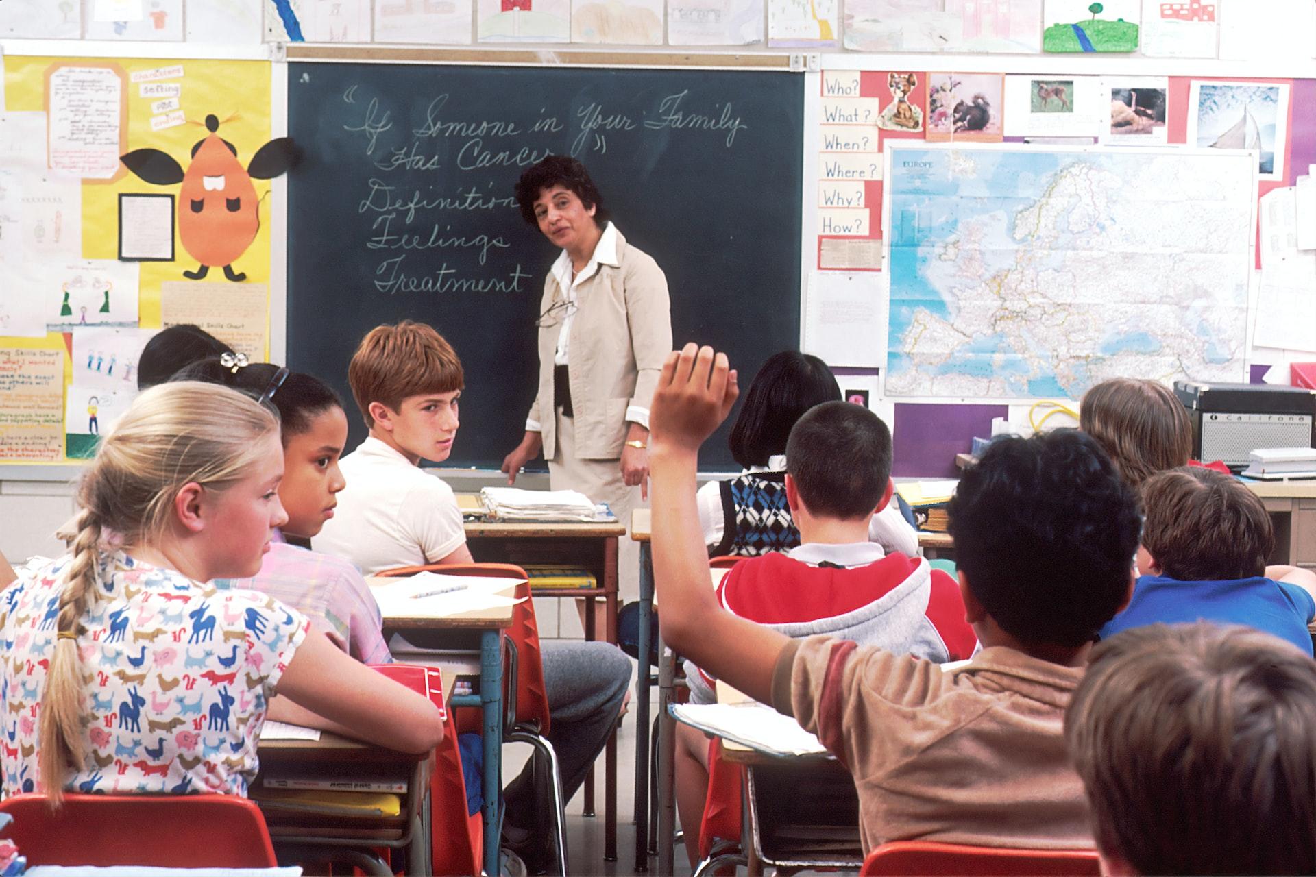 A bright, cheerful classroom with students sitting at their desks and the teacher at the board and one student raising his hand while others turn toward him.