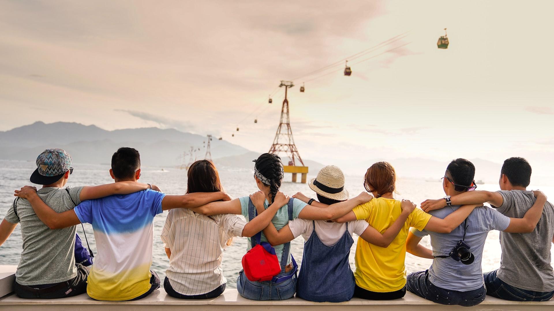 A line of people with their backs to the camera, sitting arm-in-arm on a low wall at sunset.