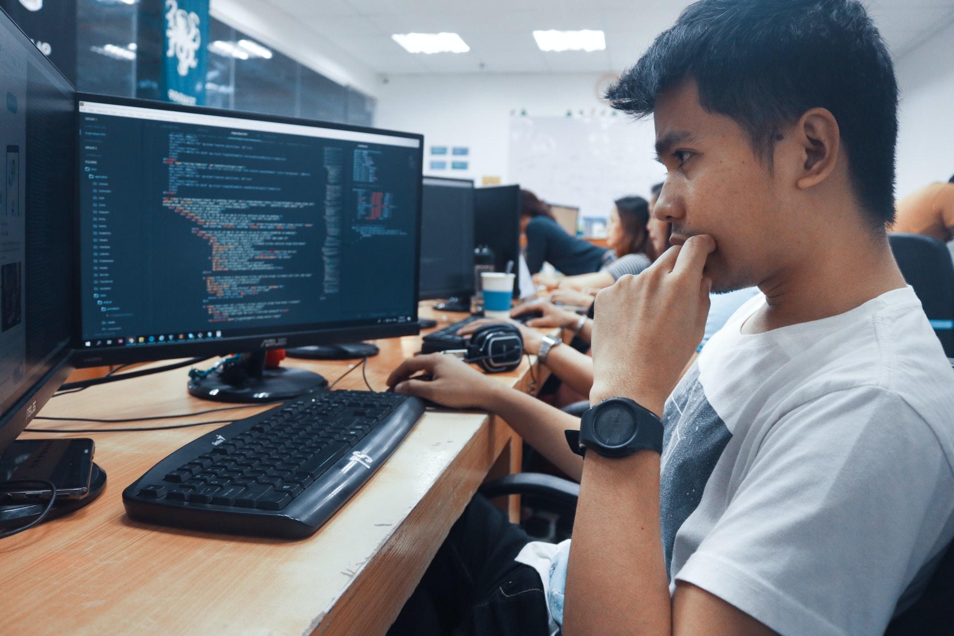 A young man sits at a wood-topped desk in front of a computer monitor looking at lines of code