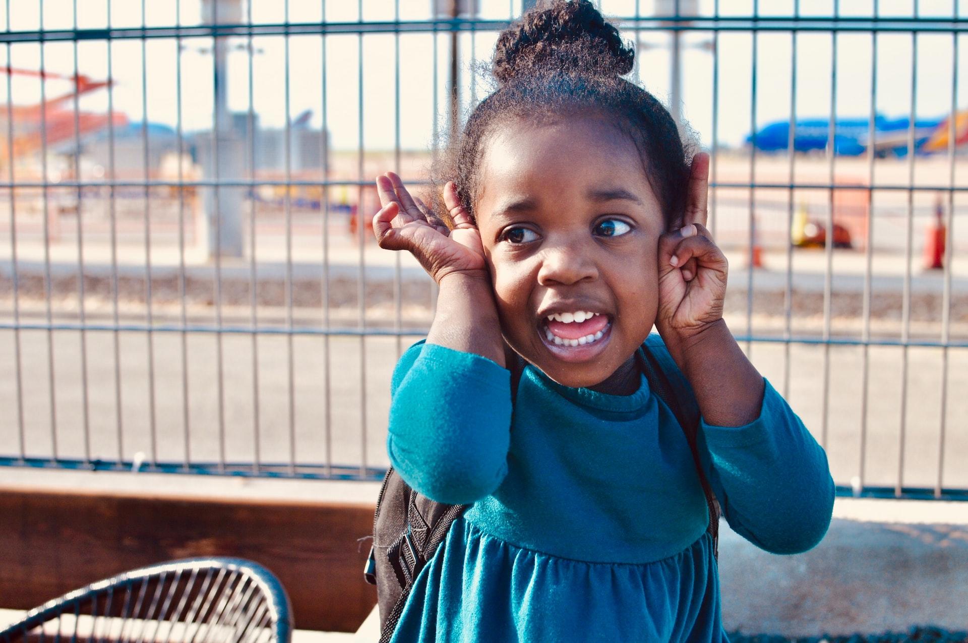 A small girl in a blue jumper with her hair piled atop her head stands outdoors, with her hands raised to her face and smiling.