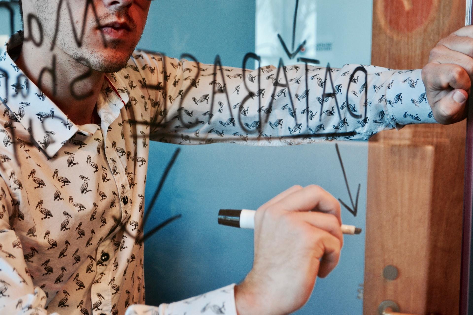 A person in a patterned shirt stands before a clear dry-erase board drafting a plan for database access