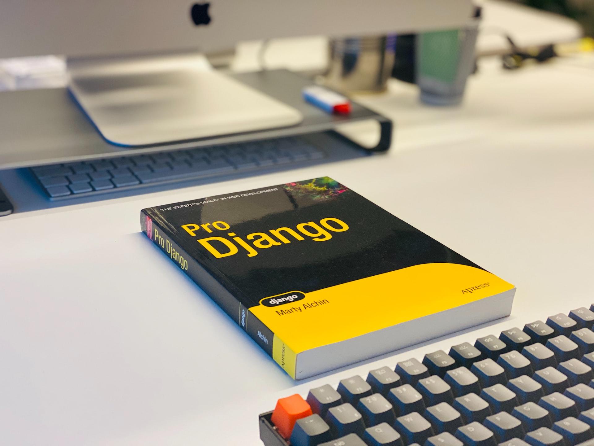 A white tabletop bearing a yellow and black Django instruction manual laying between a computer keyboard and the computer monitor's base.