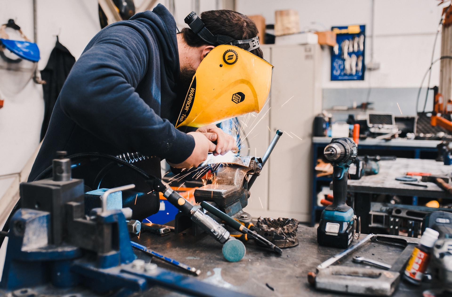 A worker in a dark coloured top wears a yellow face shield as he grinds metal on a workbench, with other workplace implements visible.