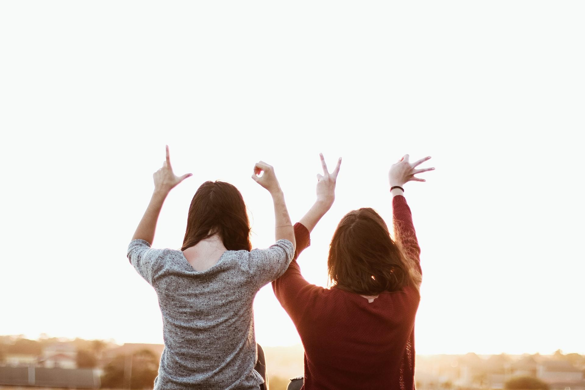 Two people with their backs to the camera, one wearing a grey top and the other, a red one, hold their hands up in the shaped to spell the word 'love'. 