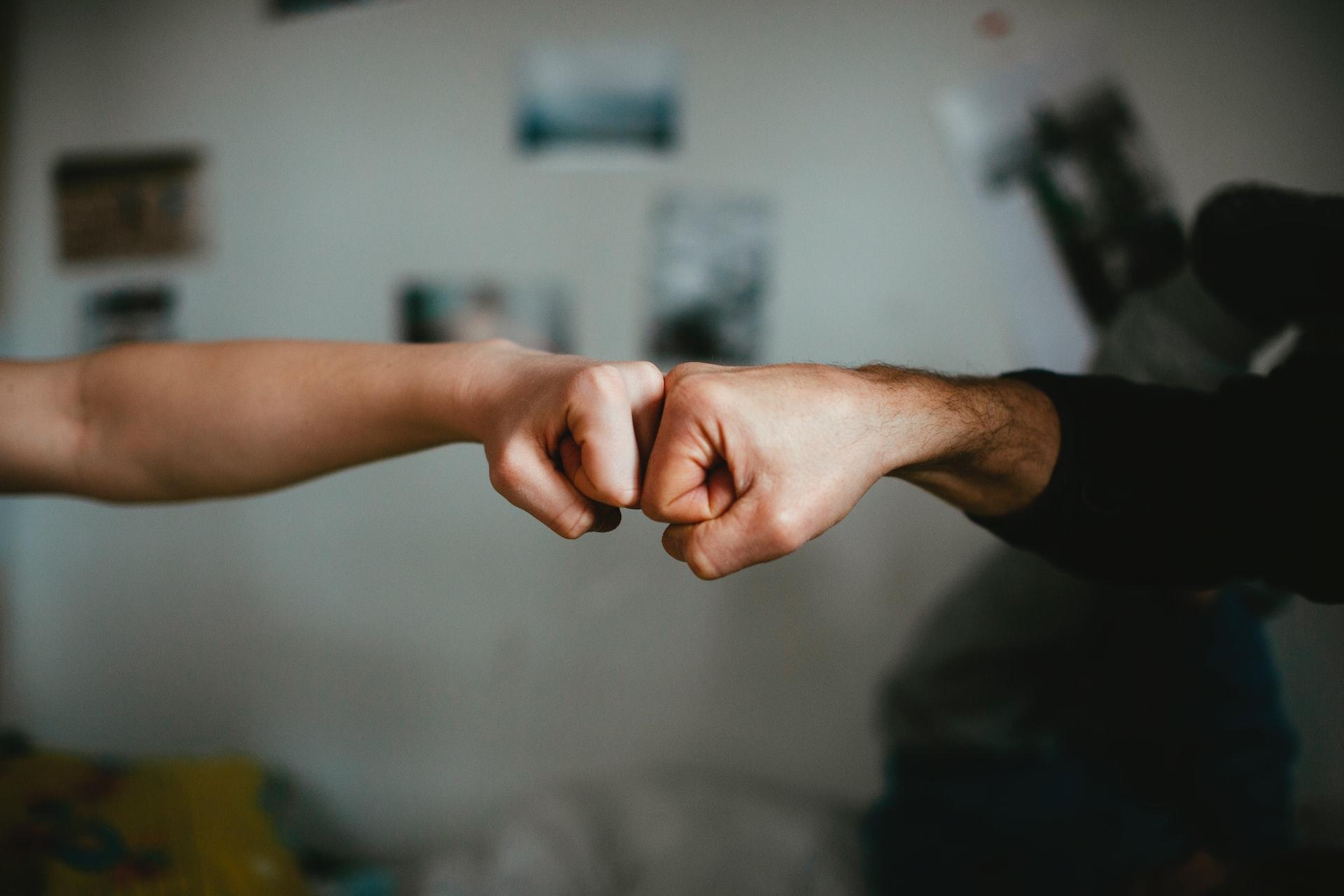 Two fists bumping in solidarity in an otherwise empty frame, with three blurry pictures hanging on the wall in the background. 