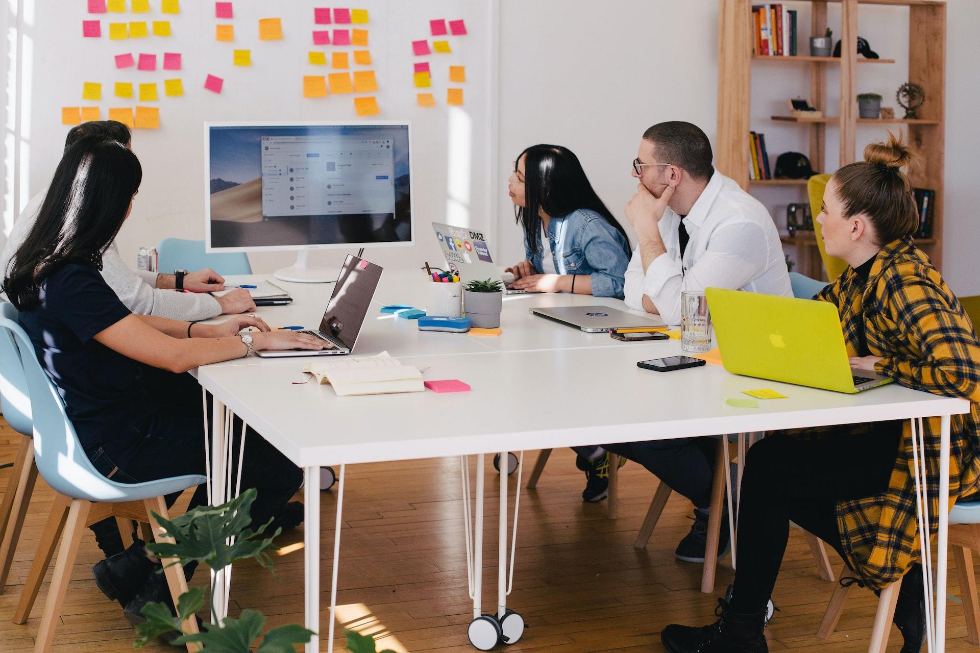 A group of people sit at a white table, focused on the computer monitor on a wall bedecked with fuchsia and orange sticky notes.