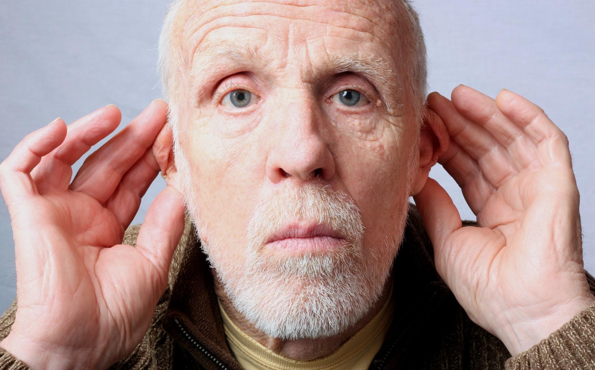 A man with a white beard stubble cups his hands around his ears with a blank expression.