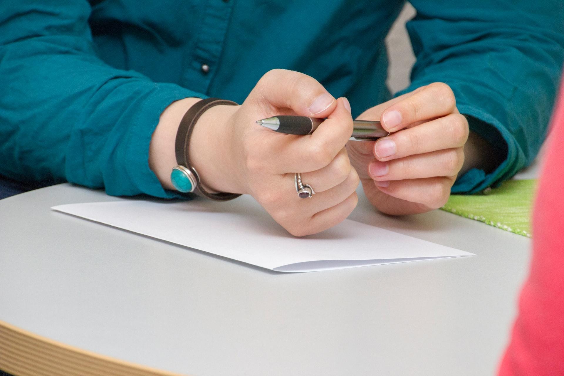 A person in a green top and wearing a bracelet sits at a table with a blank sheet of paper in front of them and a pen loosely gripped in their hand.