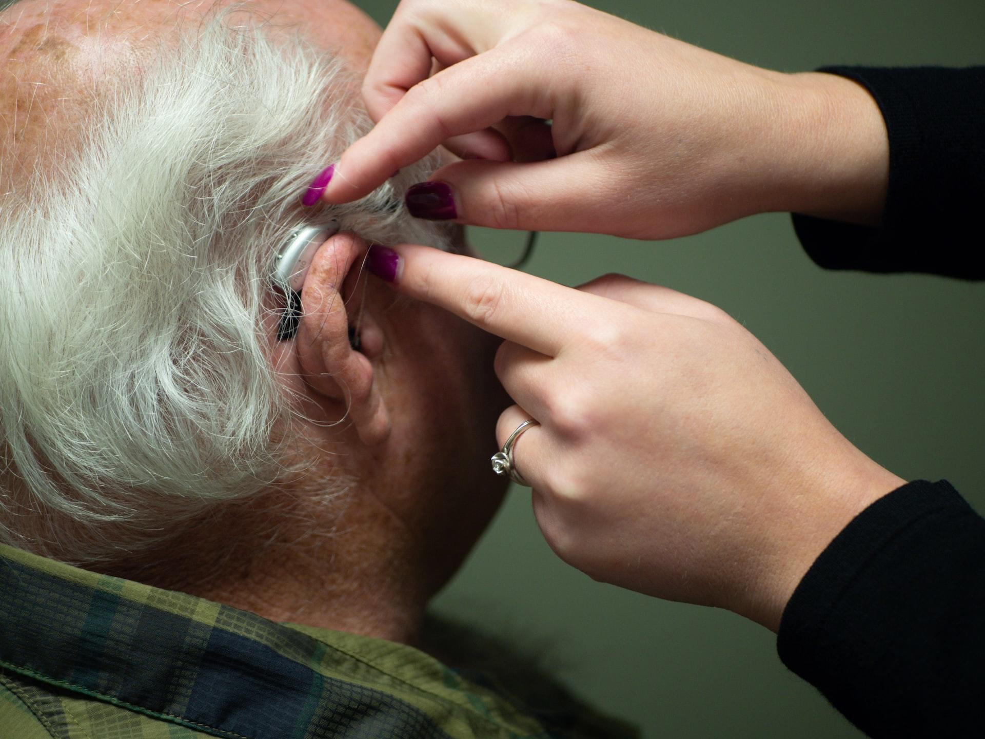 An older man with silver hair wears a hearing aid, which a younger person with fingernails painted red is adjusting.