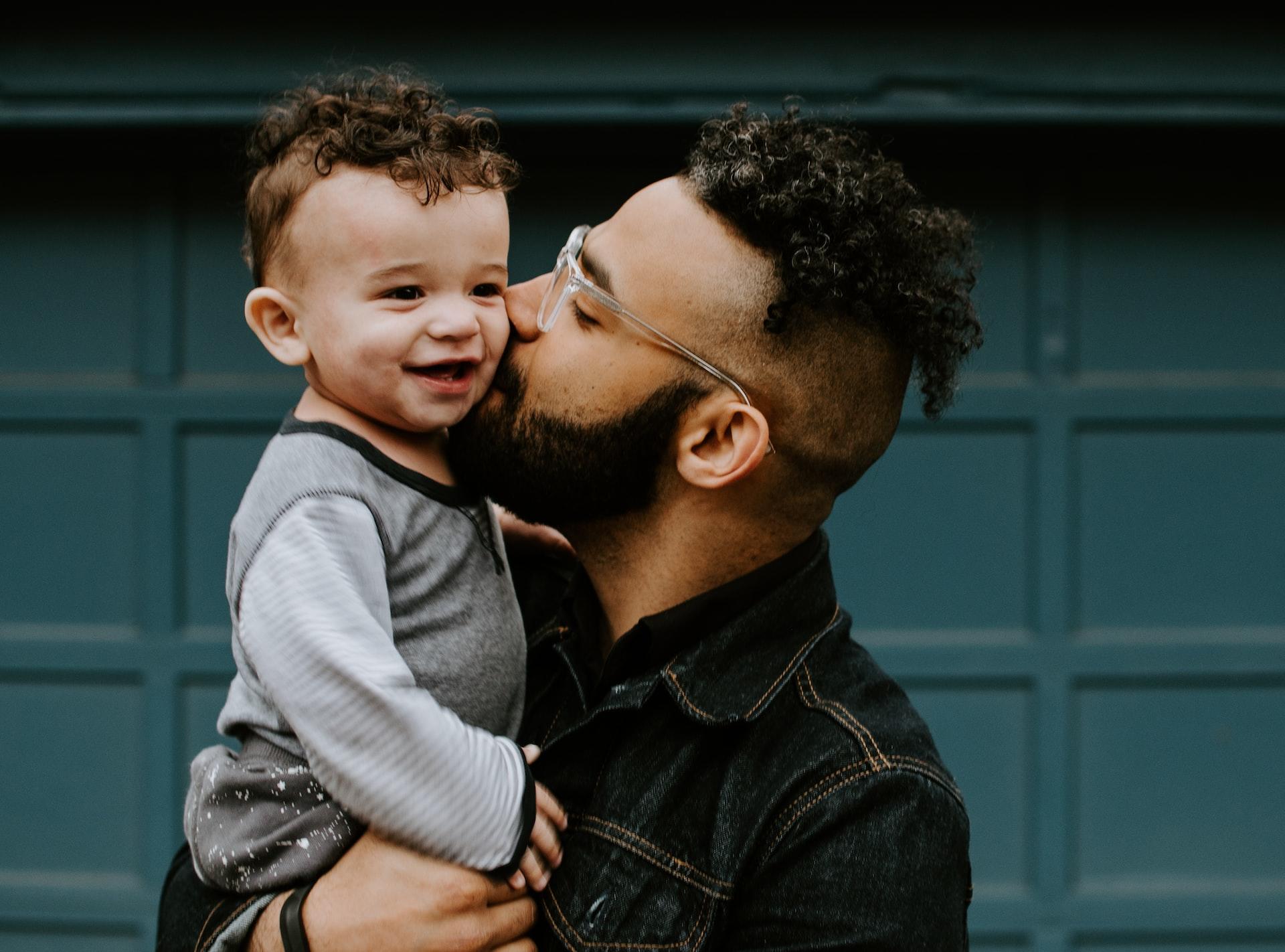 A man with glasses, wearing a dark top holds a baby wearing a light-coloured top and kisses him on the cheek as the baby smiles.