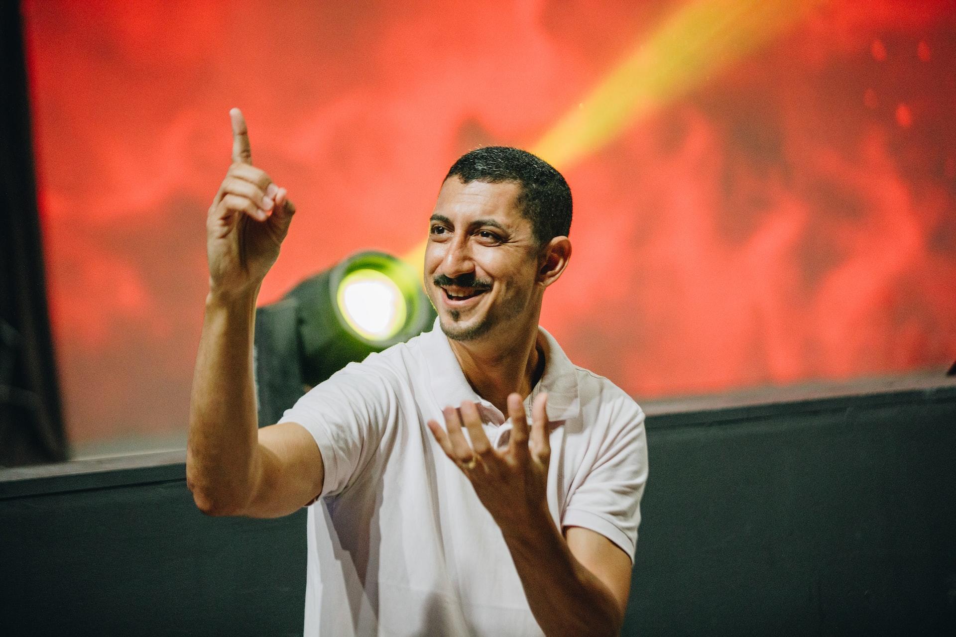 A man in a white shirt stands against an orange backdrop with a yellow spotlight shining in front of it, smiling and using sign language.