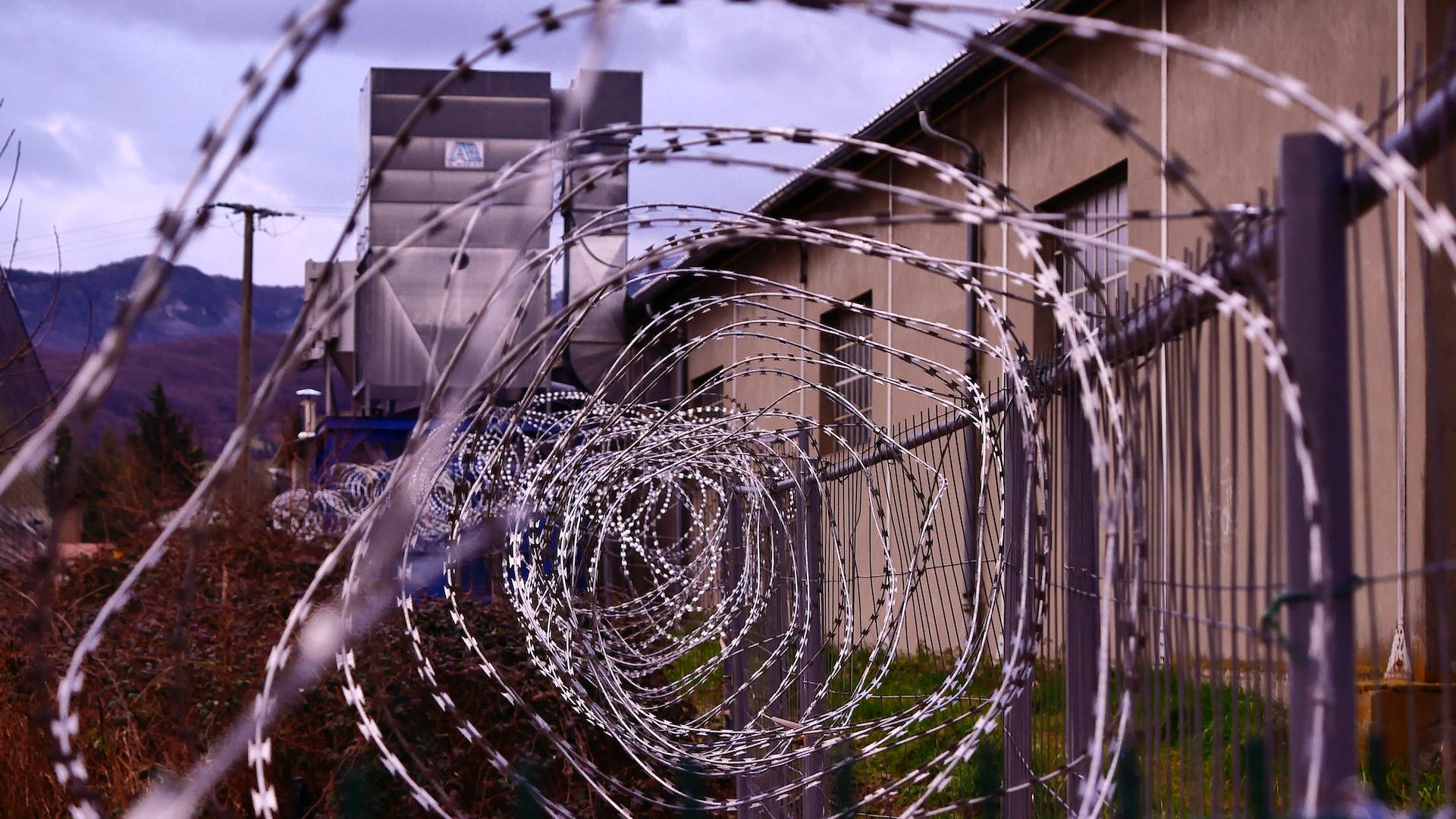 A coil of concertina wire spread atop a fence in front of a squat, tan building with a tower visible in the background.