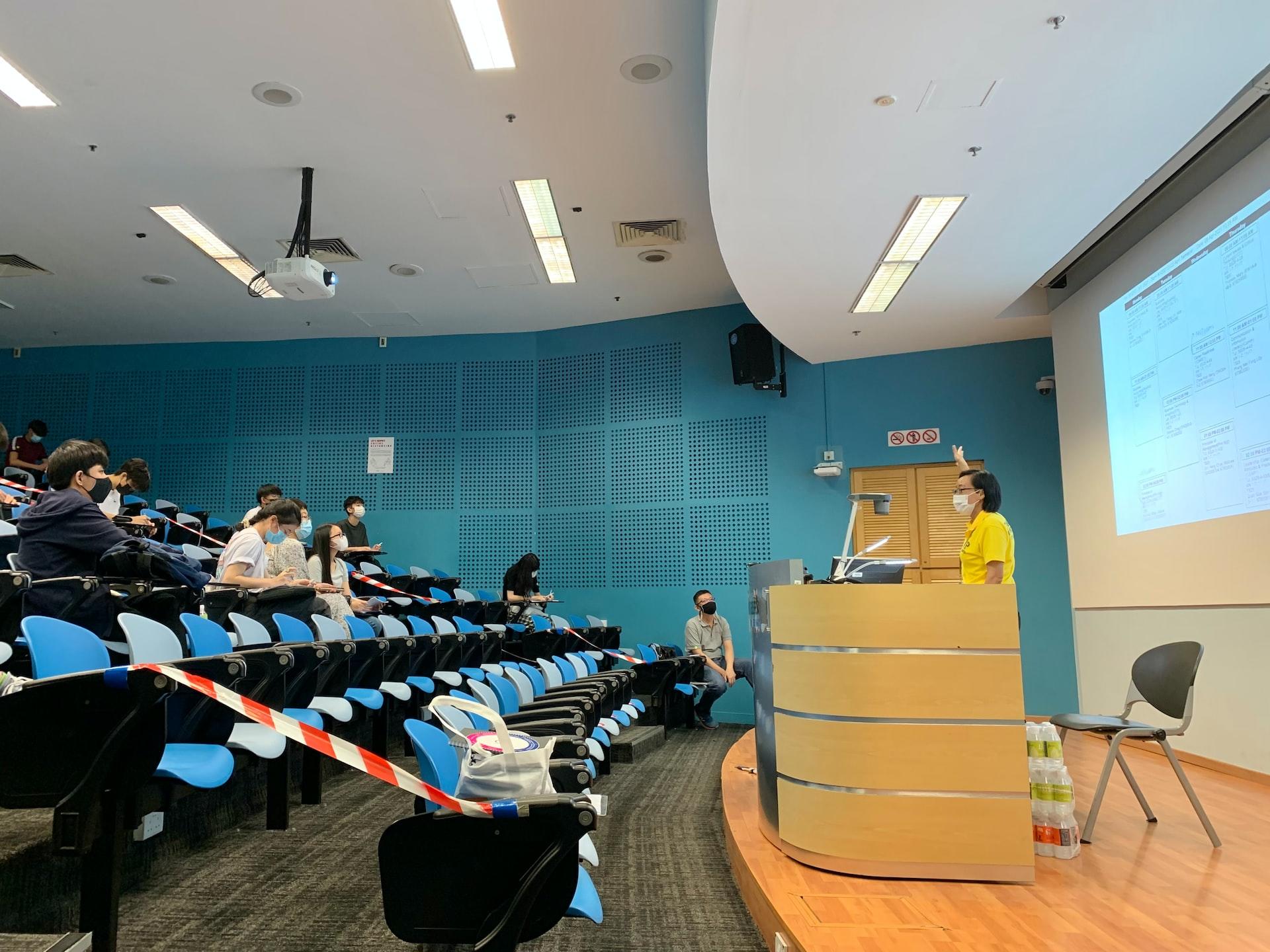A university lecture hall with aquamarine walls and blue seats facing a light-coloured dais and projection screen.