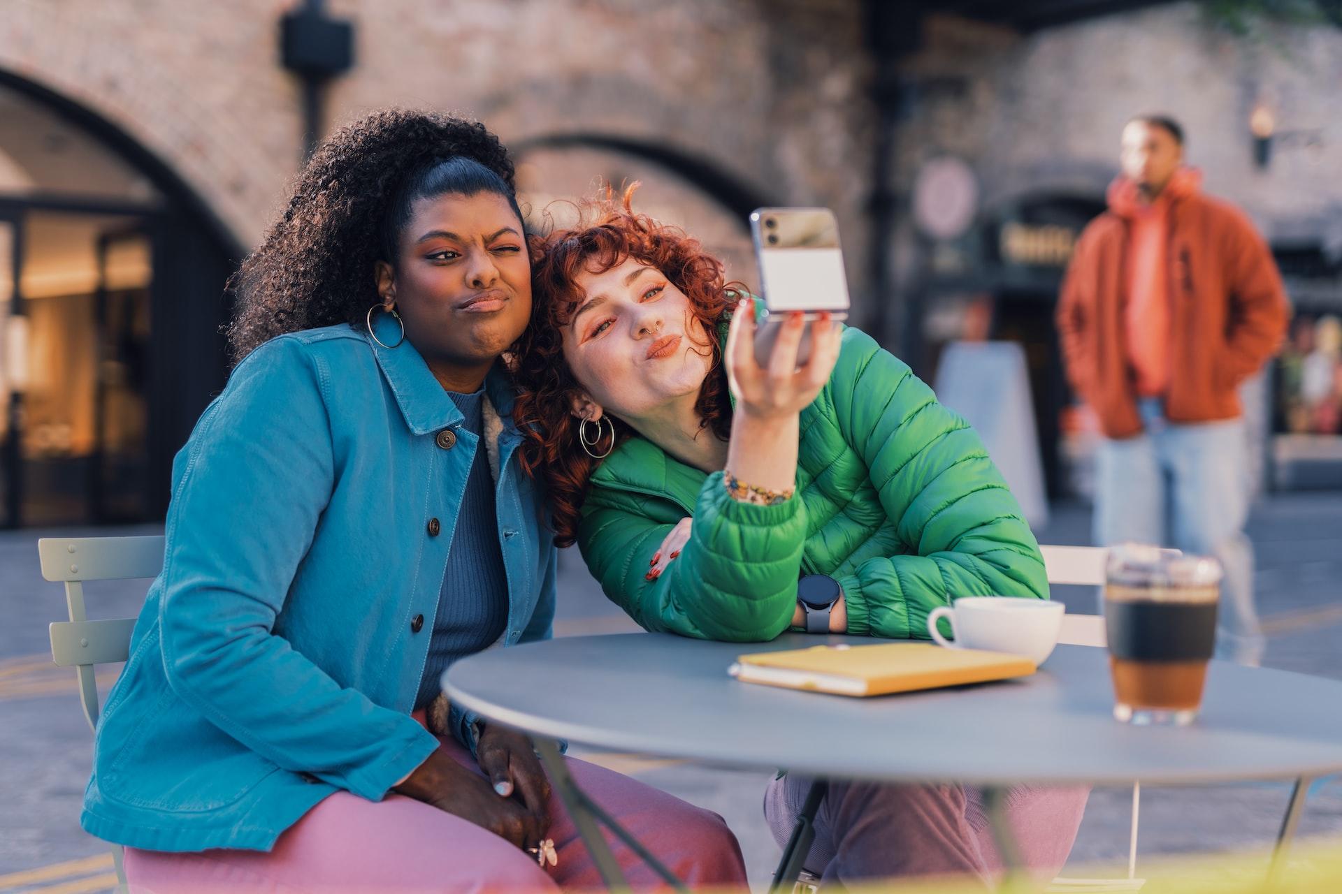 Two women, one in a blue coat and the other in a green puffy jacket, sit at a table in an outdoor space, posing for a selfie.