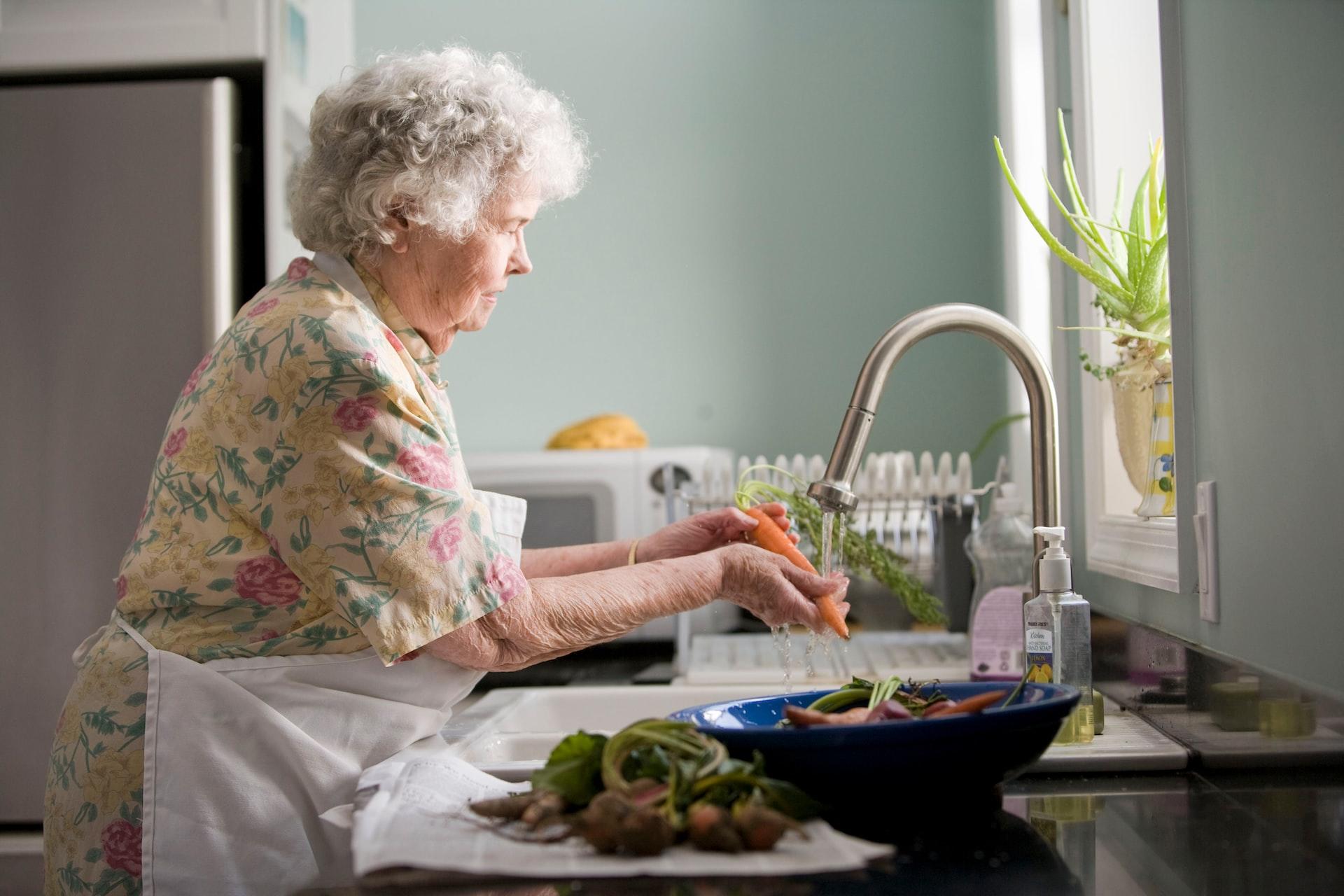 An elderly woman wears an white apron over her flowered shirt stands at the kitchen sink, washing carrots.