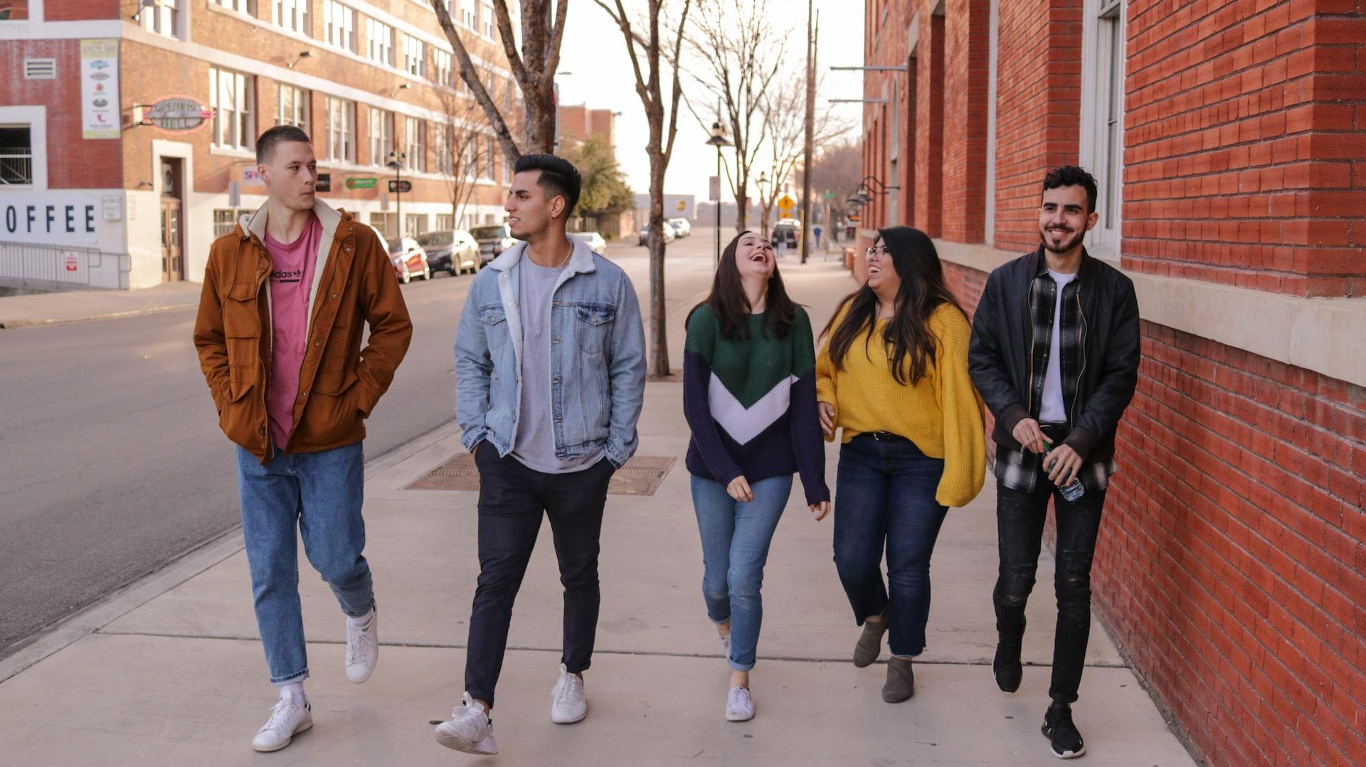 A group of young adults wearing autumn jackets walk along the sidewalk, past denuded trees, laugh together.
