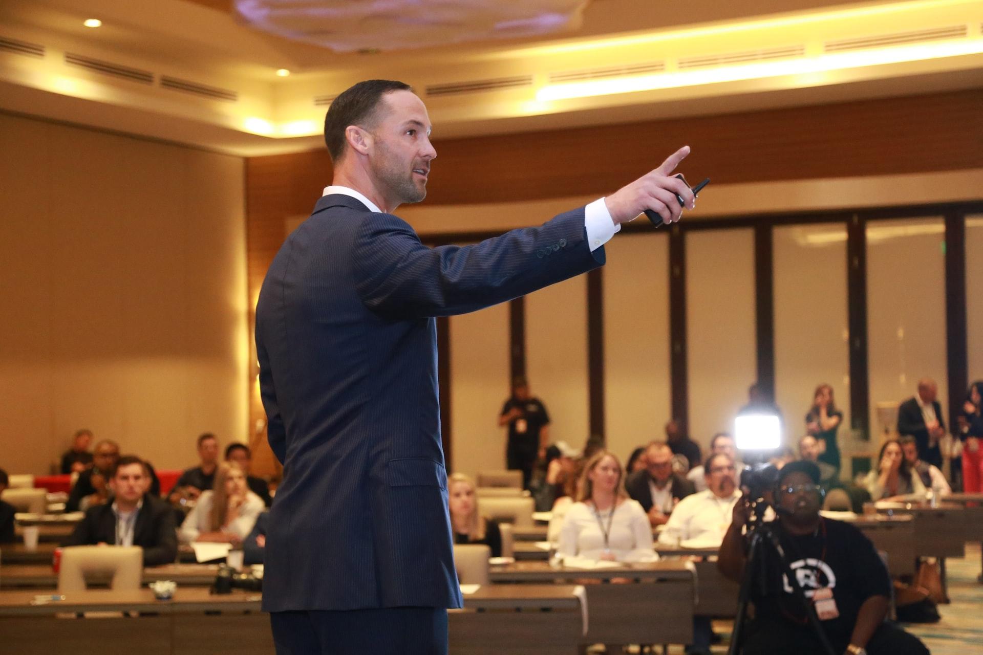 A man in a business suit stands in a courtroom, pointing at something while people in the gallery look on.