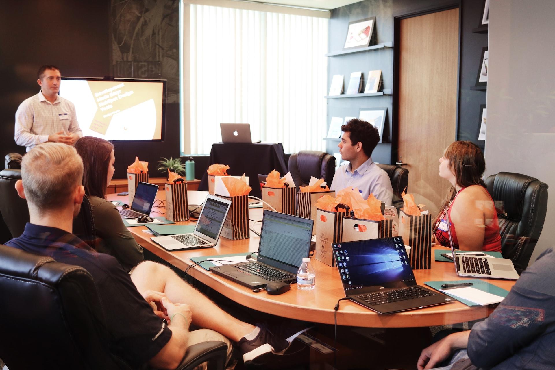 Five people sitting at a table laden with open laptop computers and gift bags listen to a person in a white shirt standing by a display monitor.