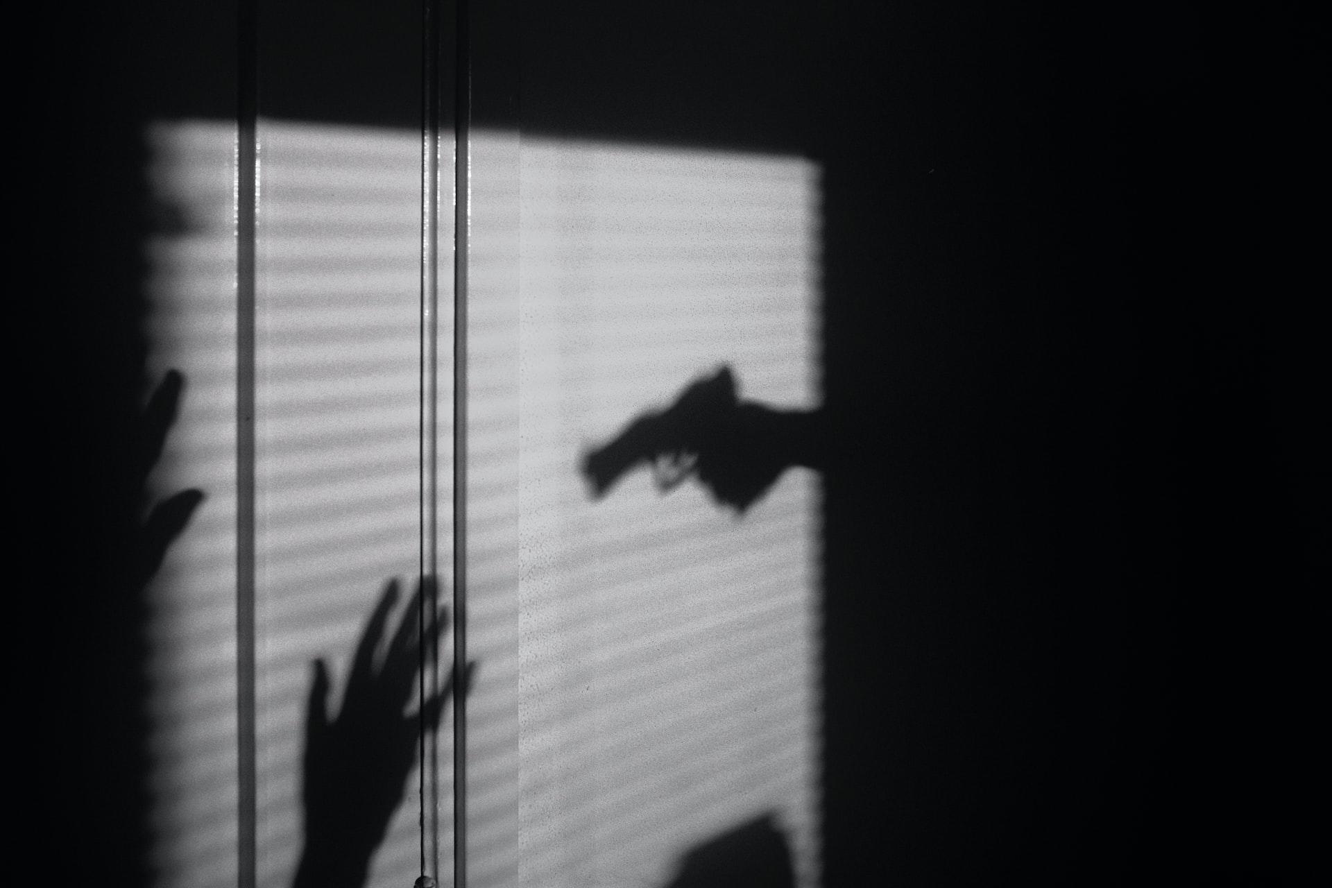 A greyscale image of a domestic violence situation with a shadow of a gun pointed at an upraised hand, set against a window with shades covering it.
