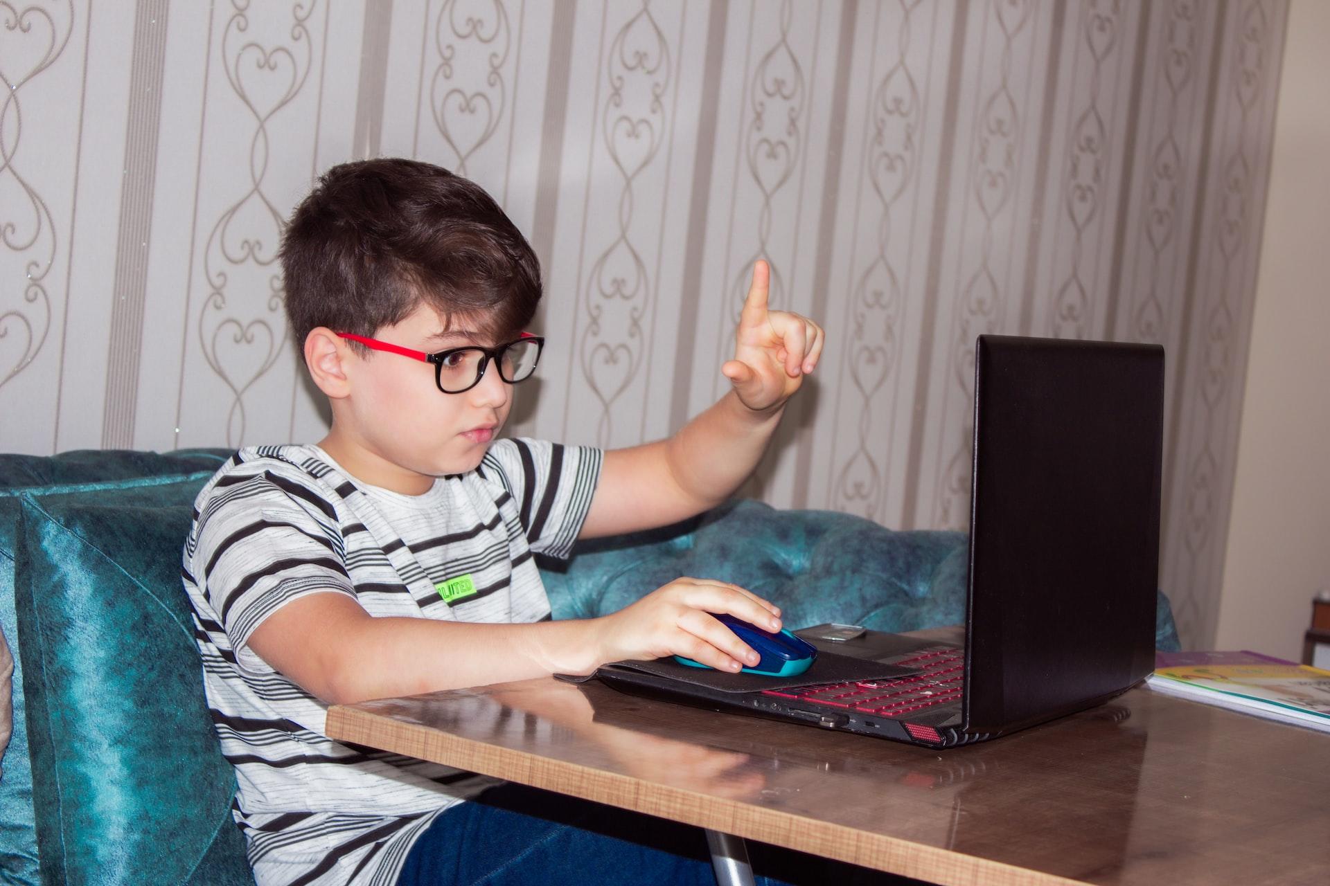 A boy wearing a striped shirt and glasses with red branches sits in front of an open laptop holding up one hand while the other hand rests on his computer's mouse.