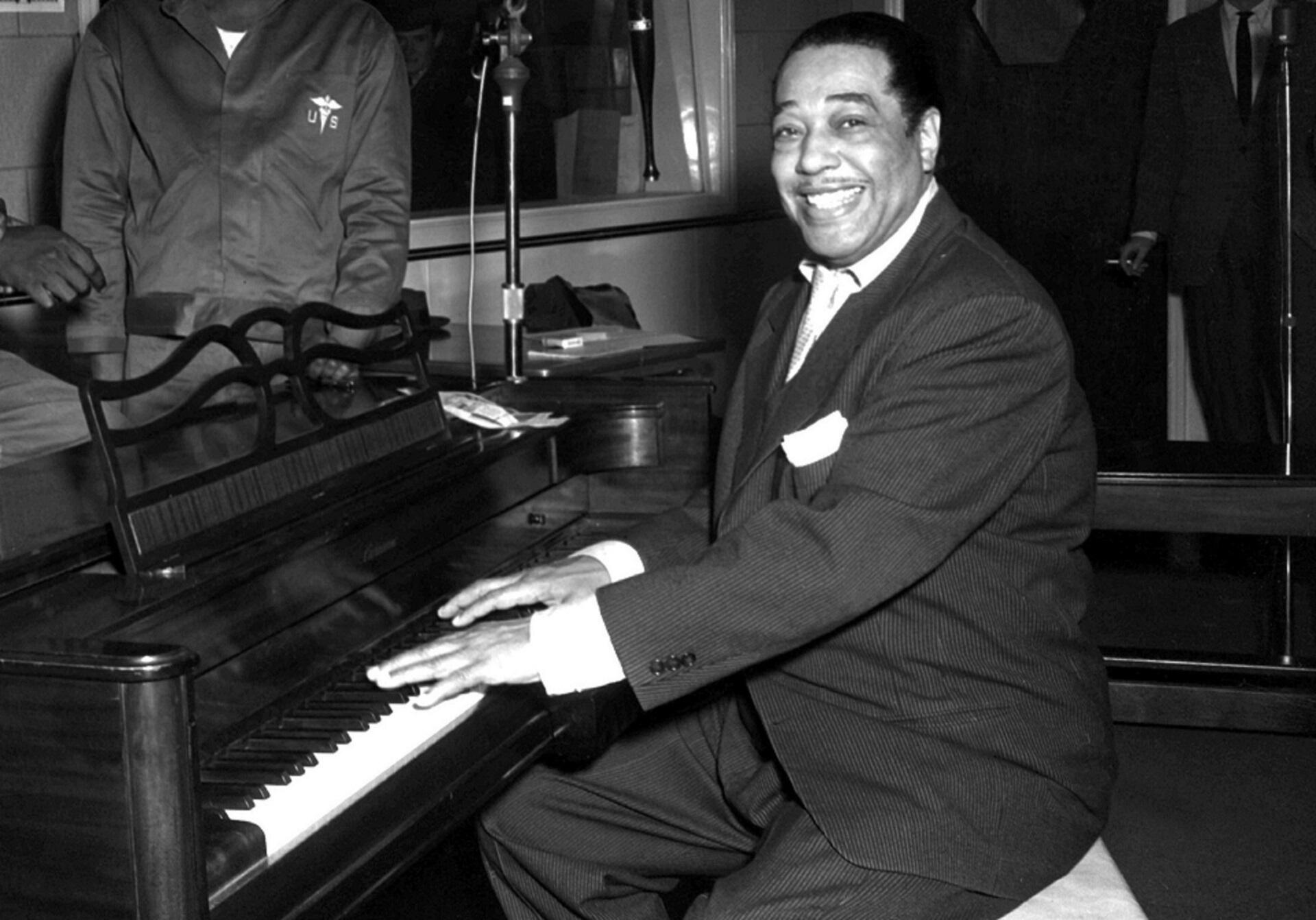 A man in a suit and tie sits with his hands on a piano keyboard, beaming and looking at the camera.