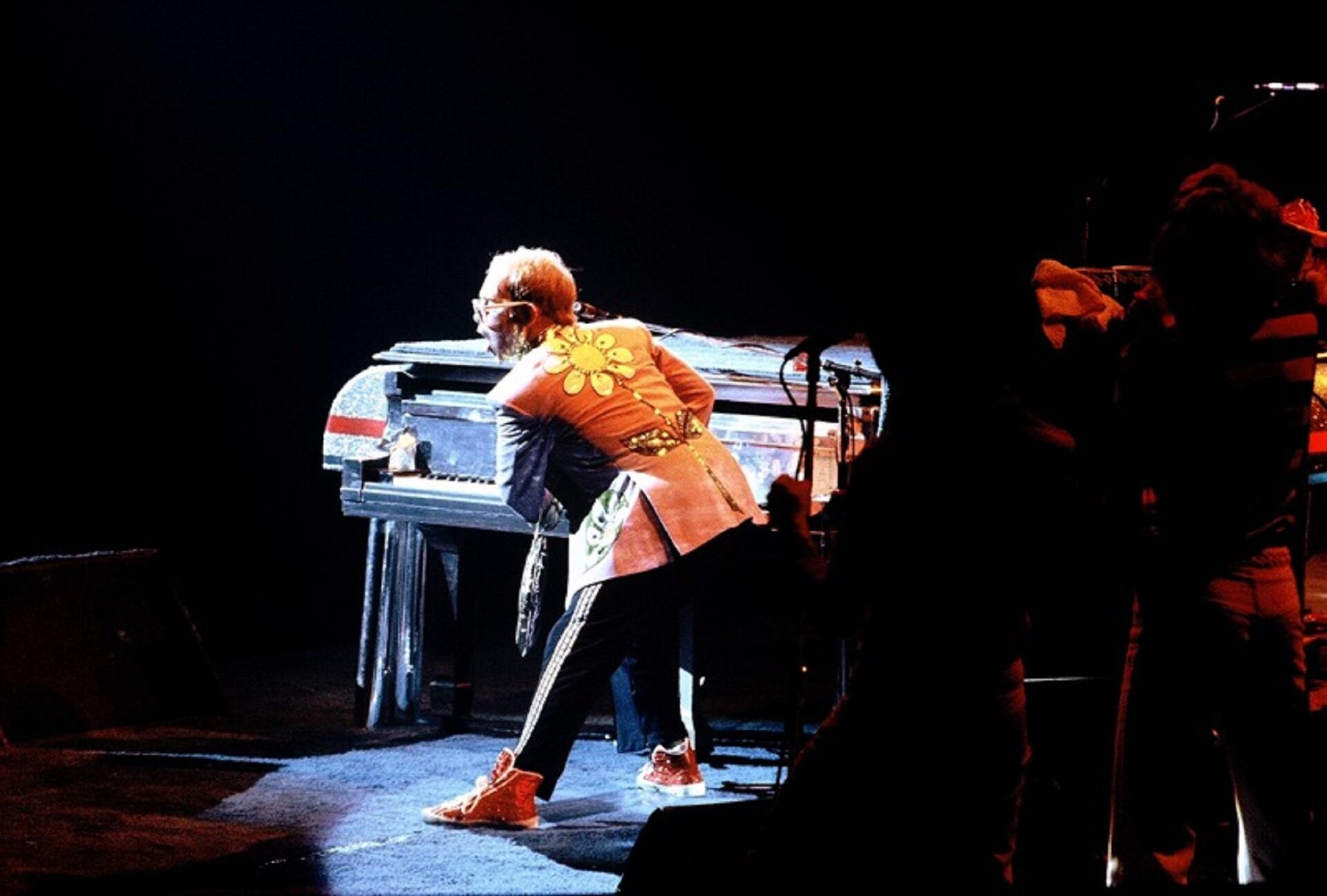 A person strikes a strange pose in front of a piano on stage while wearing a yellow coat with a large flower on the back of it.
