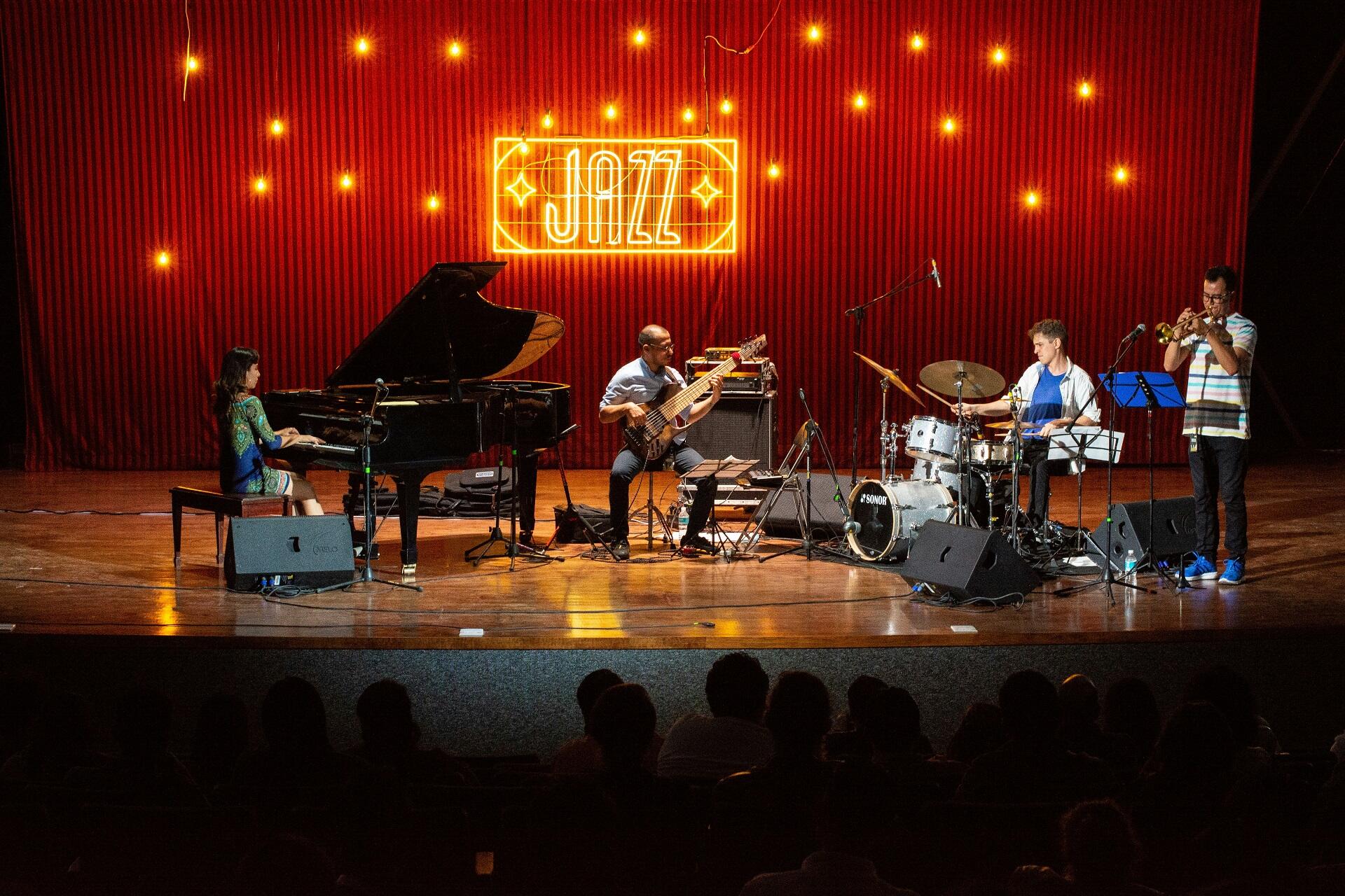 A group of musicians on stage with their instruments, in front of a red curtain with yellow stars and a lit-up 'jazz' sign on it.