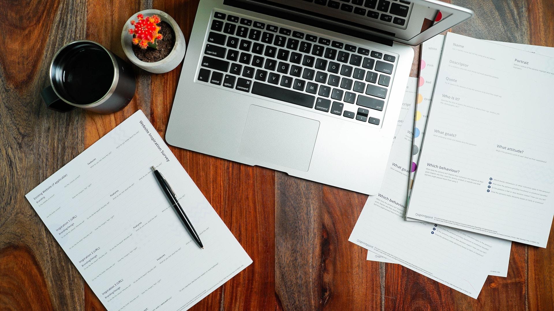 A silver laptop with black keyboard is open, next to a notepad with a black pen laying across it, a small orange plant in a white ceramic planter and a pile of papers. 