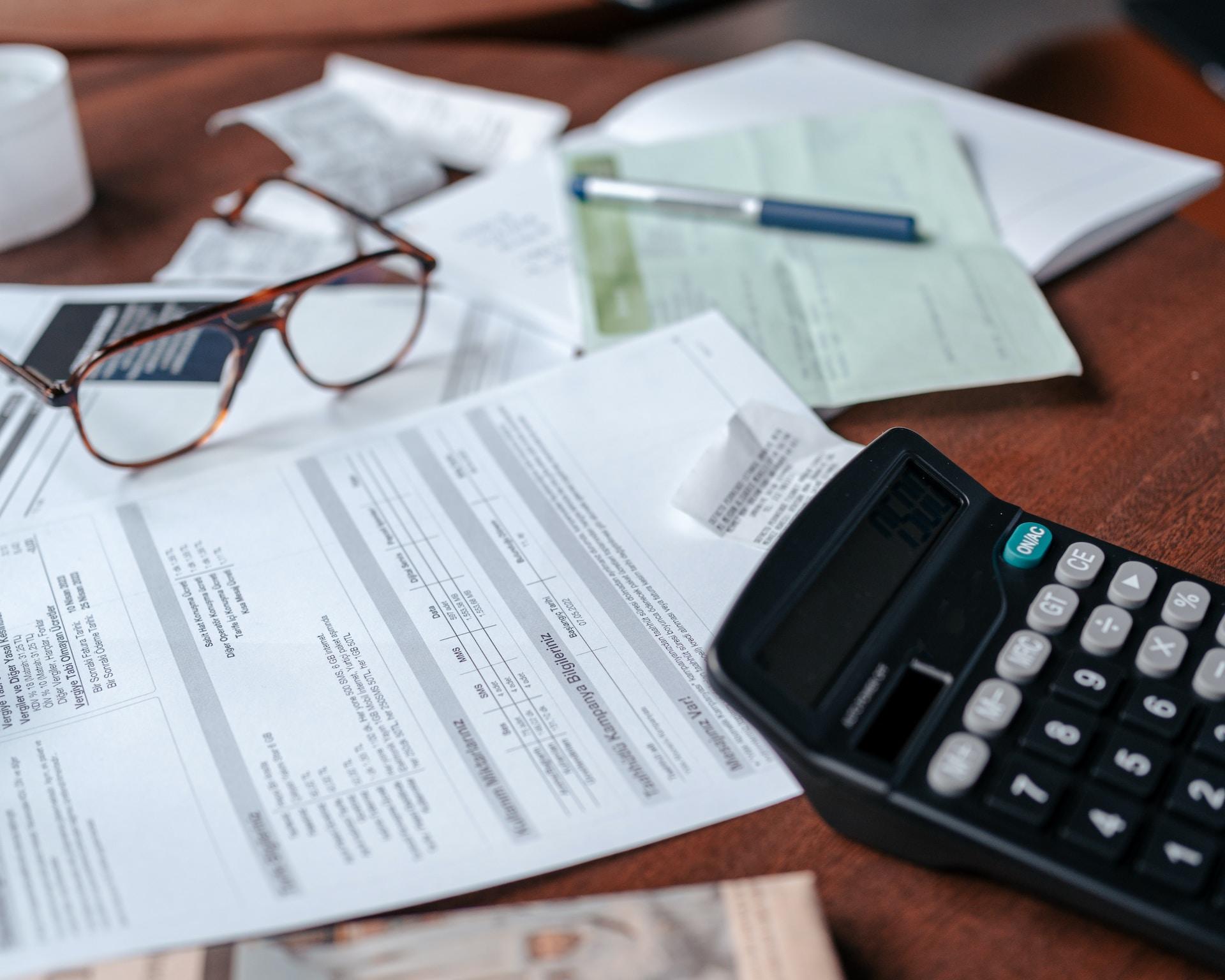 A collection of formal-looking papers scattered on a wood table, with a pen and a set of eyeglasses resting on them and a calculator with a black case and grey and black buttons nearby.