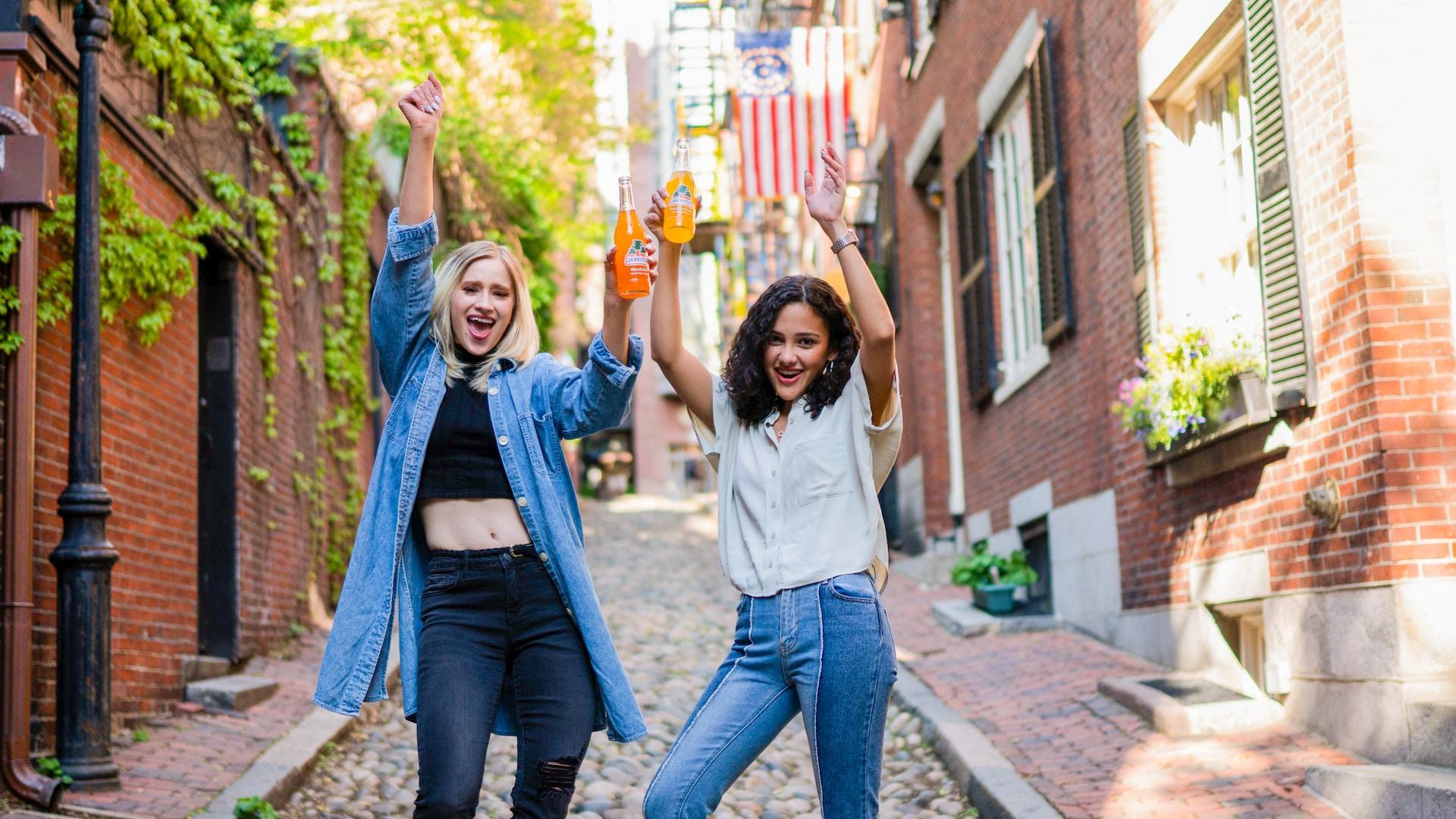Two young women wearing casual clothing strike a pose on a cobblestoned street while holding orange soda drinks aloft and smiling.