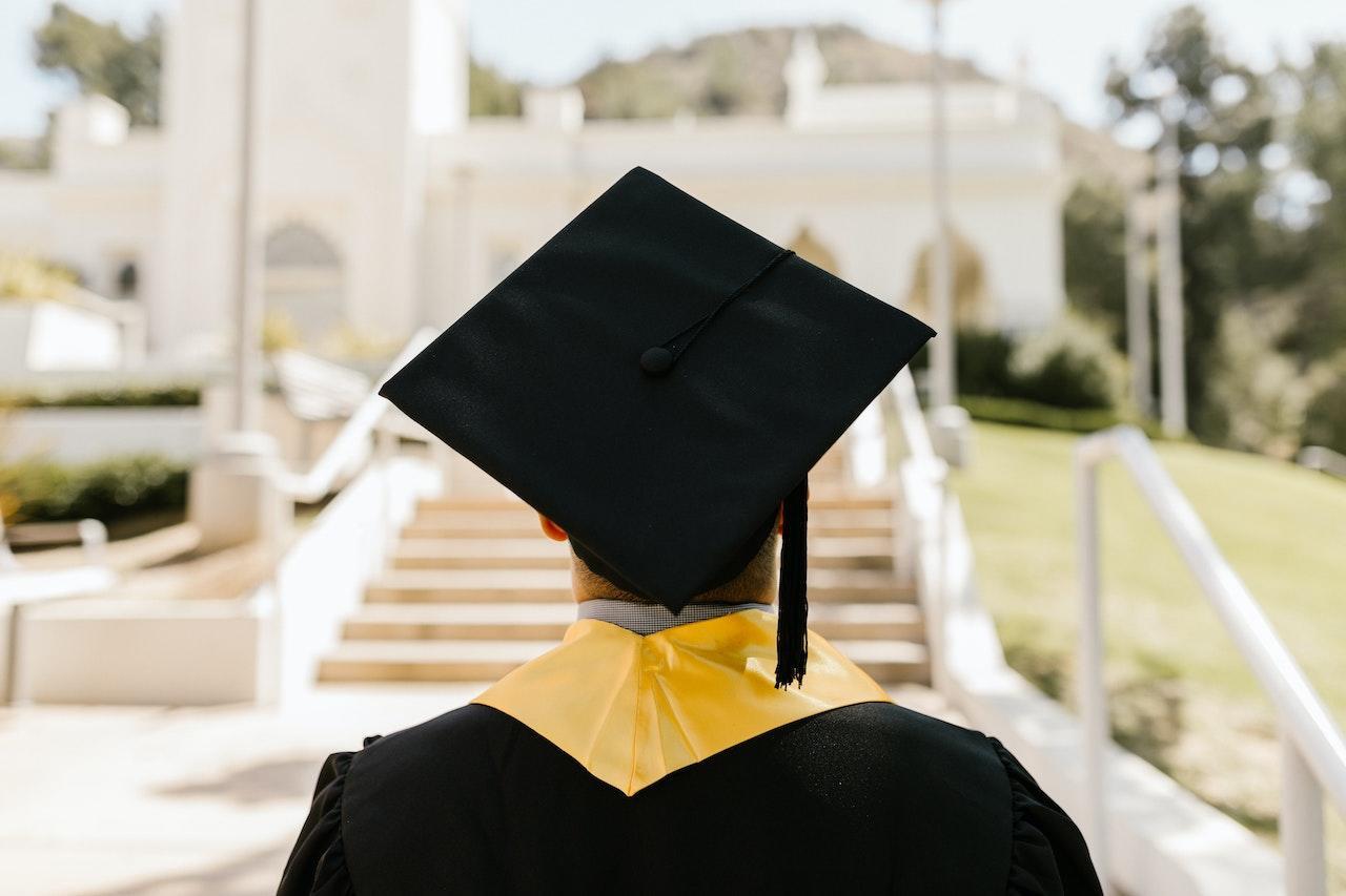 A student in a graduation cap and gown stands facing away from the camera looking towards a sunlit historic marble staircace outdoors