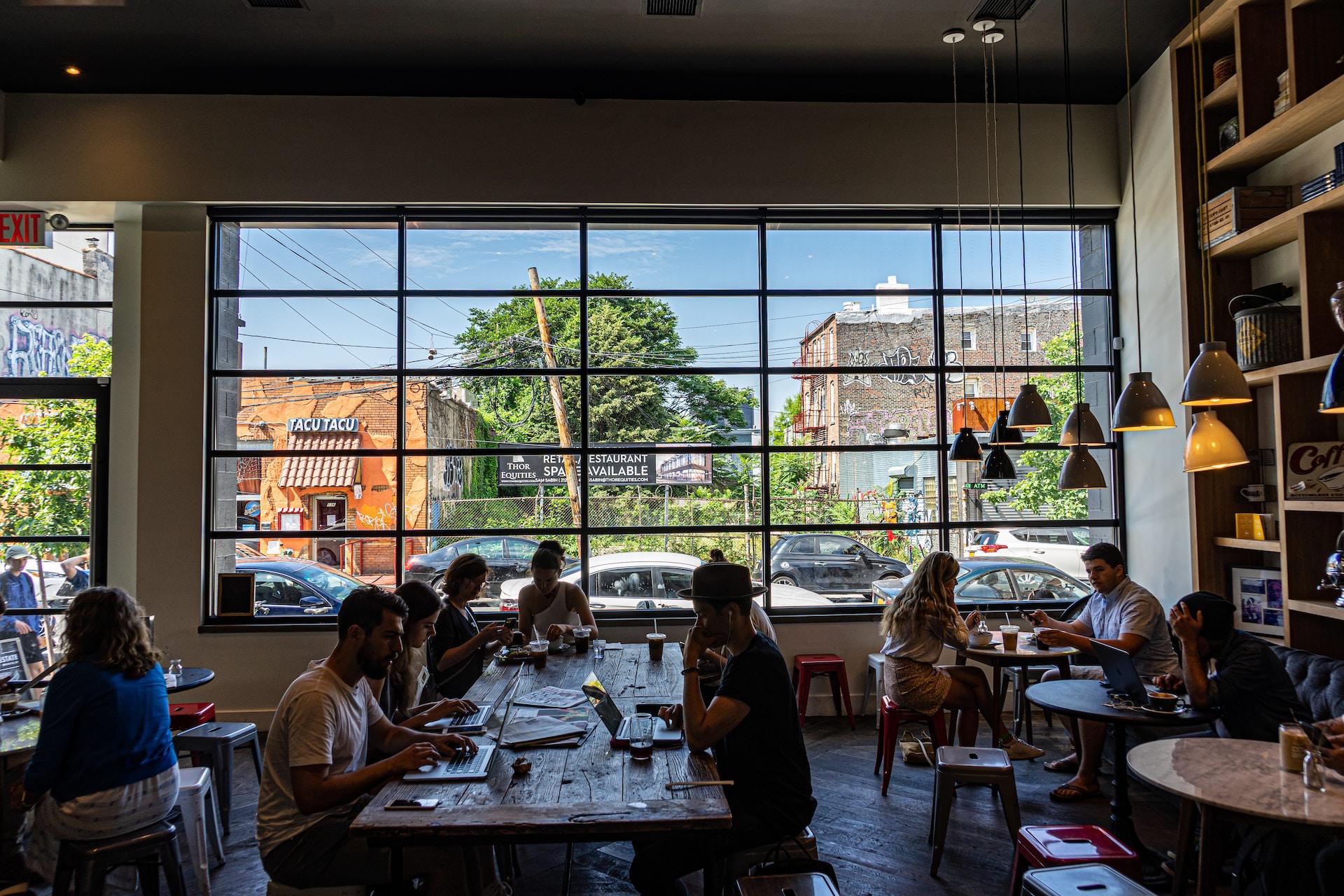 A wide-angle view of a large room that has an expansive view onto the street and fine weather outside. Several people sit at a large table laden with beverages and work materials; others sit at satellite tables scattered around the room.