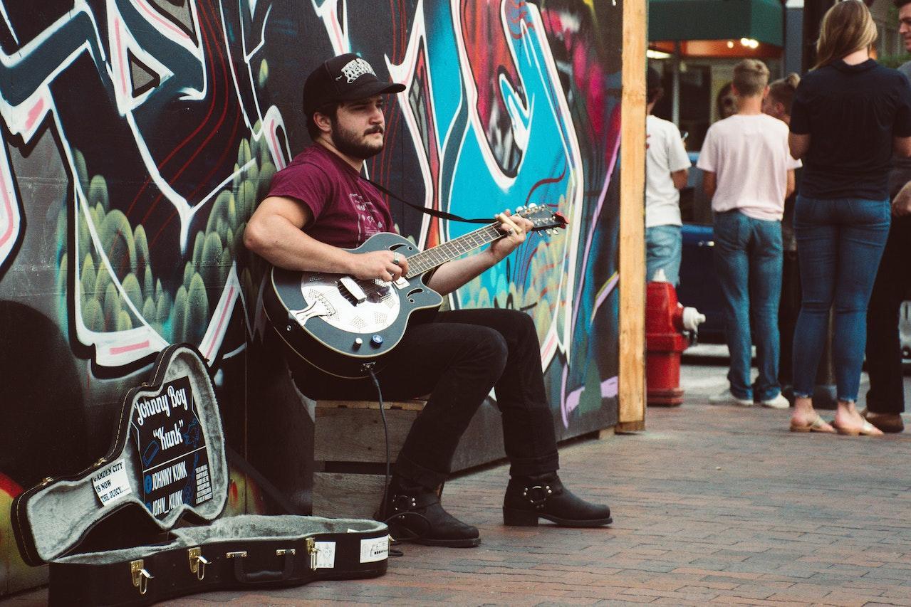 A man sits outside on a wood crate in front of a brick wall with a graffiti mural busking with a black and white guitar.
