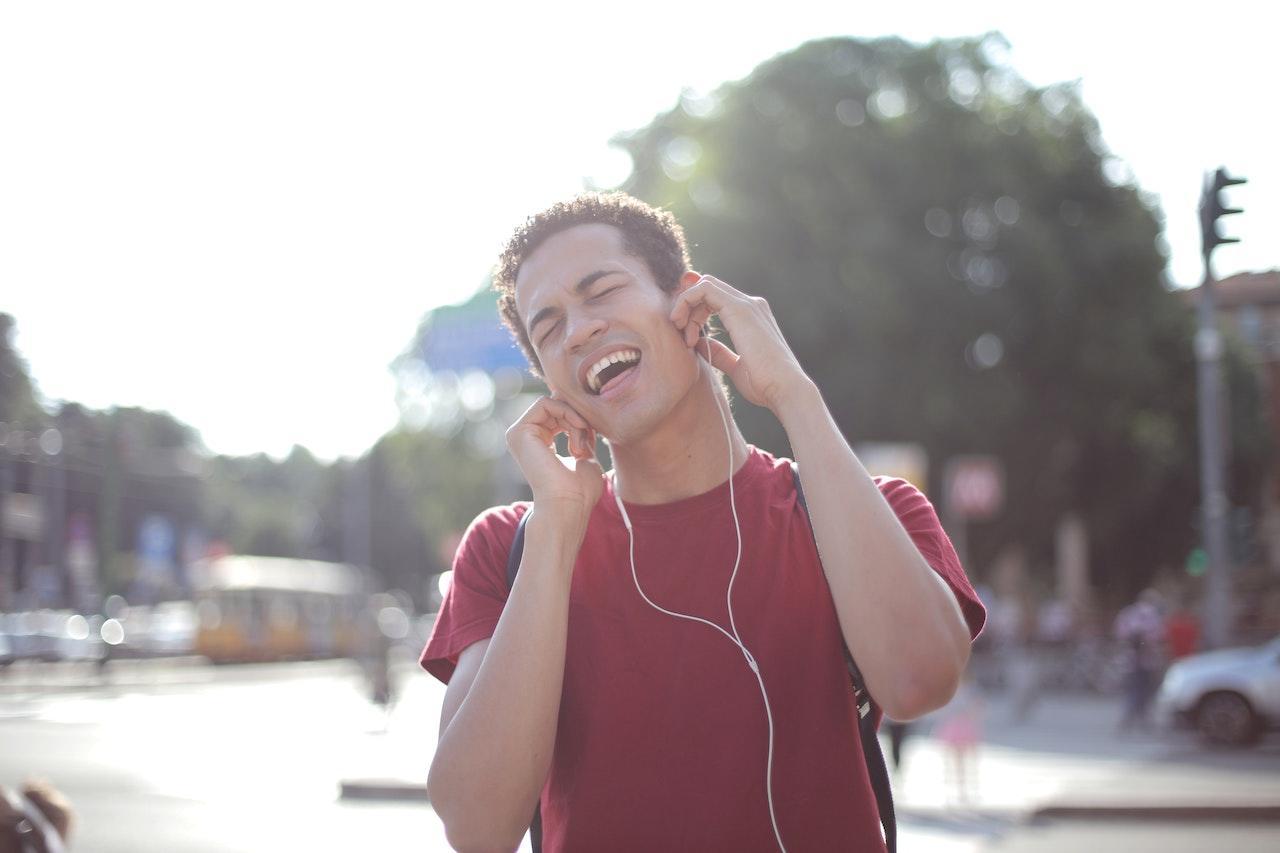 A man in a red t-shirt outside sings along with the song on his earbuds.
