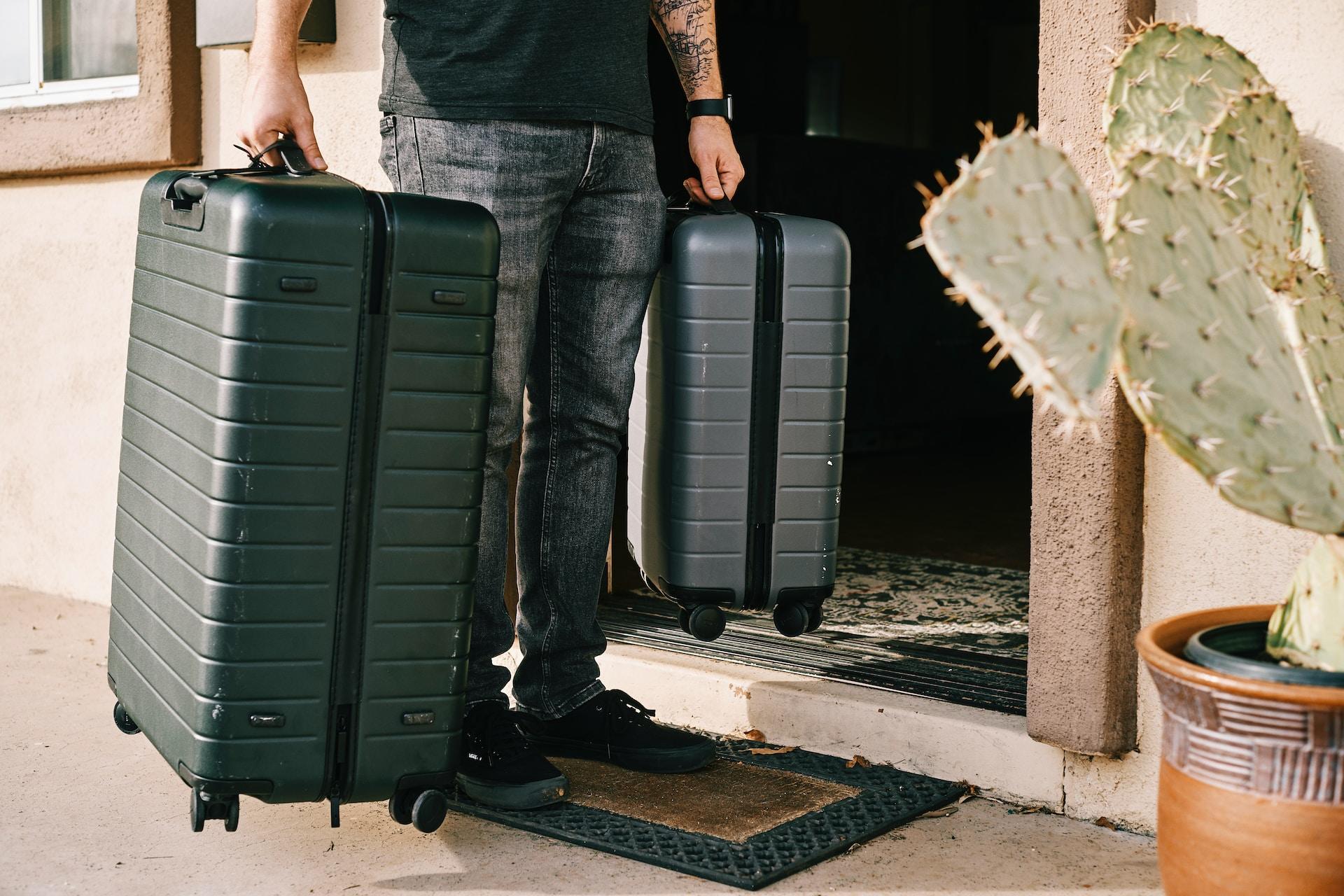 A person wearing jeans and a black shirt stands outside in front of an open door, in the sunshine, holding two black suitcases,