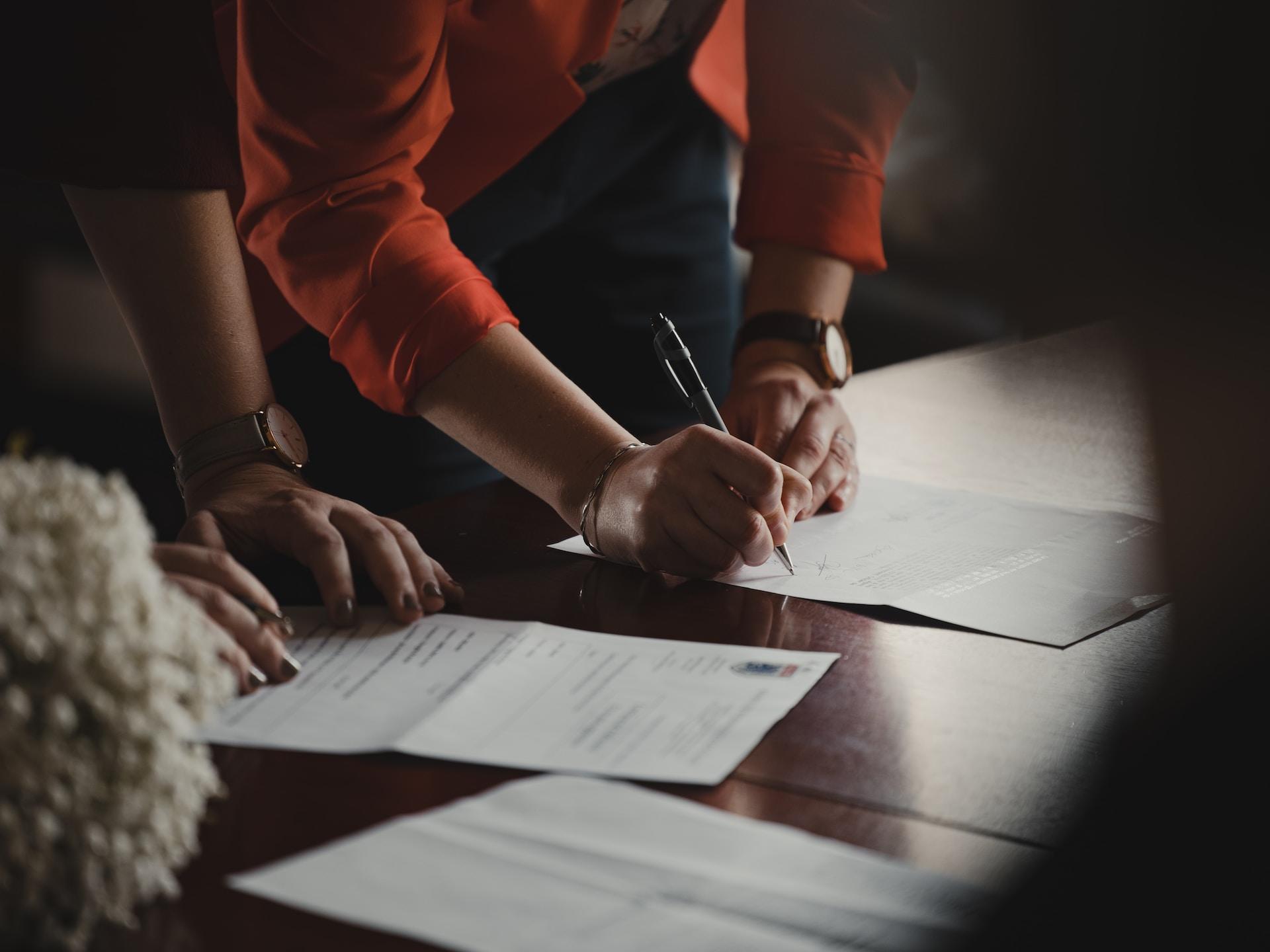 A person in an orange top signs off on paperwork while another person with fingernails painted black stands ready to hand the signer another paper to sign.