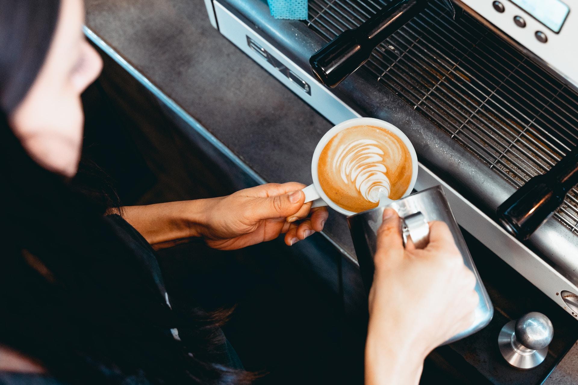 An overhead view of a person creating coffee art in the shape of a leaf behind the counter of a coffee shop.