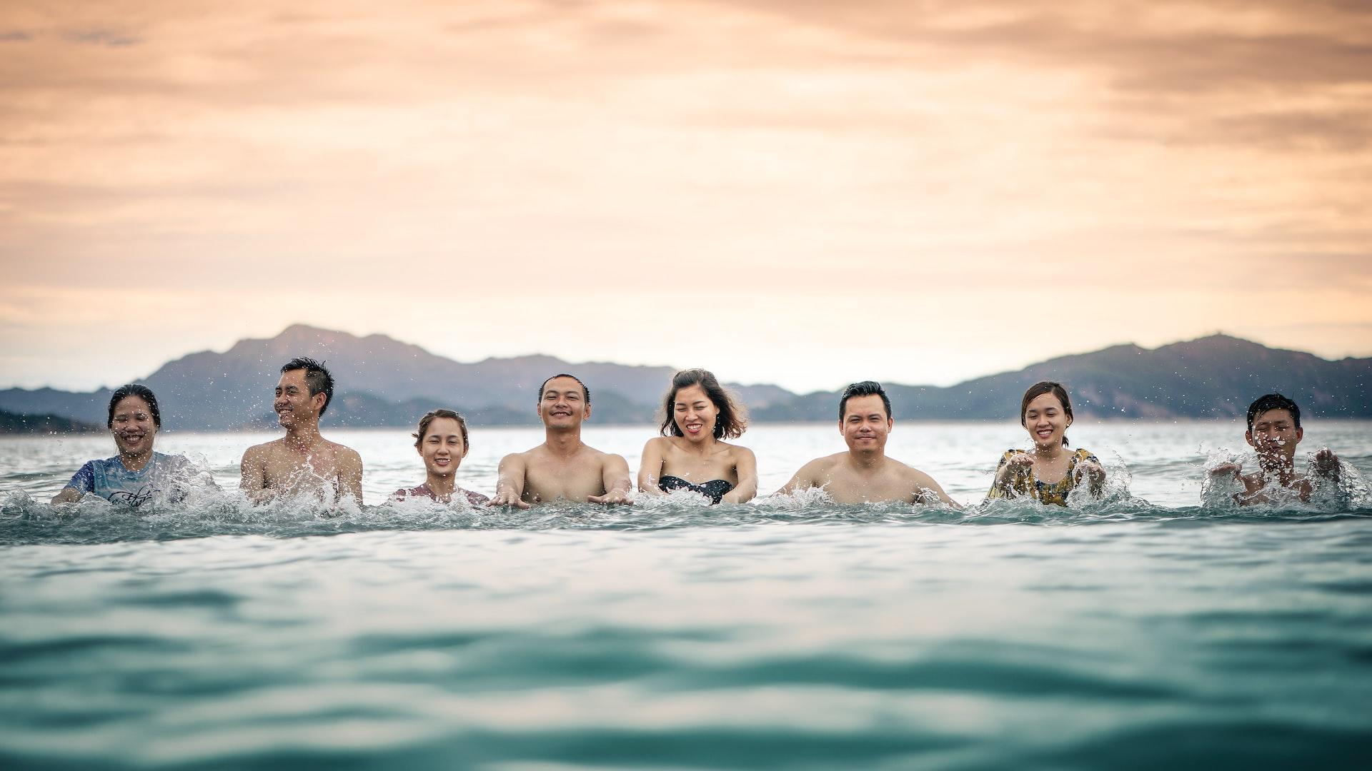 A line of young adults smiles at the camera and splashes in the ocean.