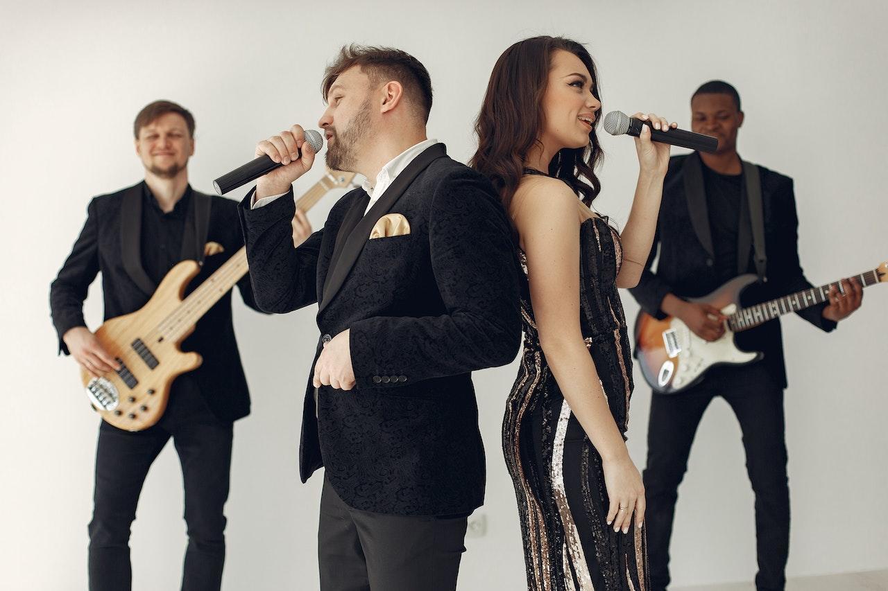 A man and a woman sing back-to-back in front of two other bandmembers who are playing guitar to either side. Everyone is wearing a mostly-black outfit with gold accents.