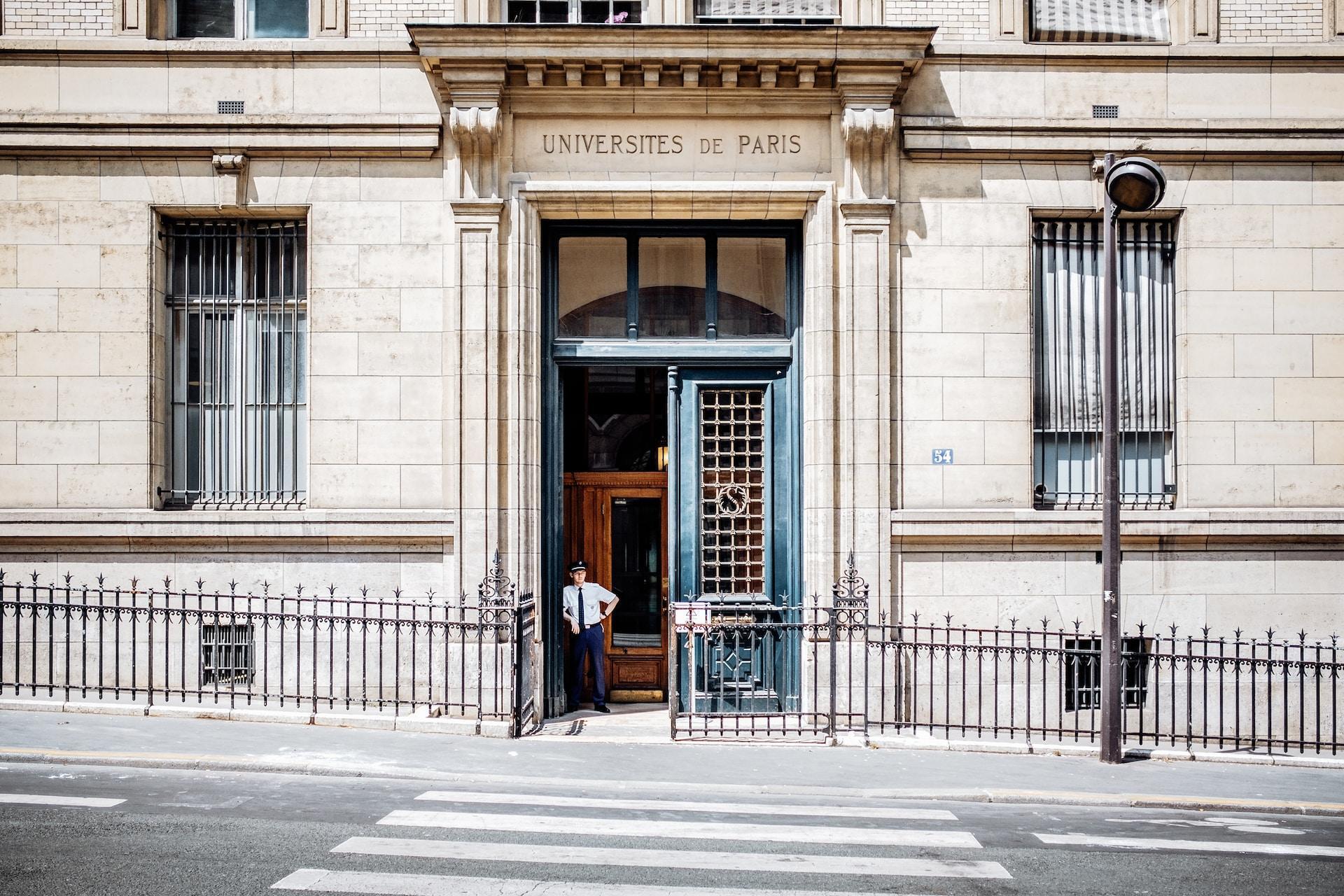 A security guard stands in front of a building titled "Universites de Paris," a formidable building made of old, large, white bricks and has a wooden door.