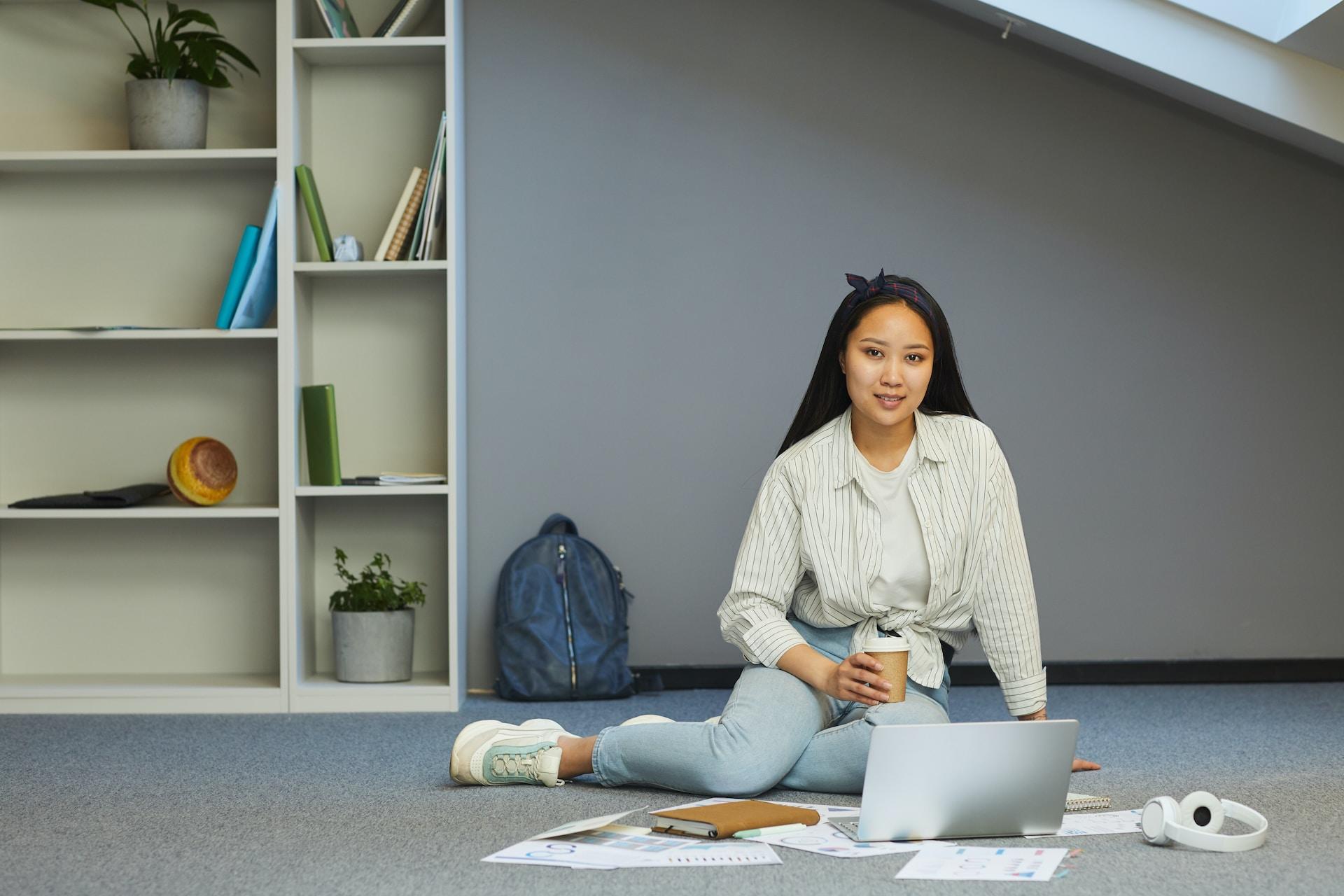 A young woman wearing a white jacket over a white top and denims sits on the floor, in front of an open laptop that is surrounded by papers, cradling a cup of drink in her right hand. 