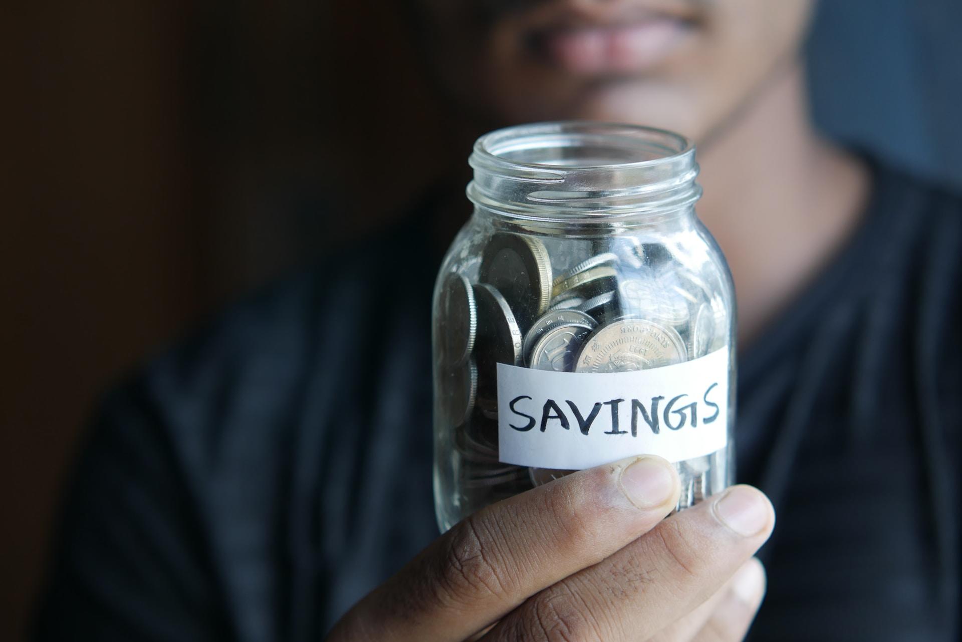 A person wearing a dark shirt holds up a small jar labelled 'savings', written in black ink on white paper. The jar is filled with coins and the person is not smiling.