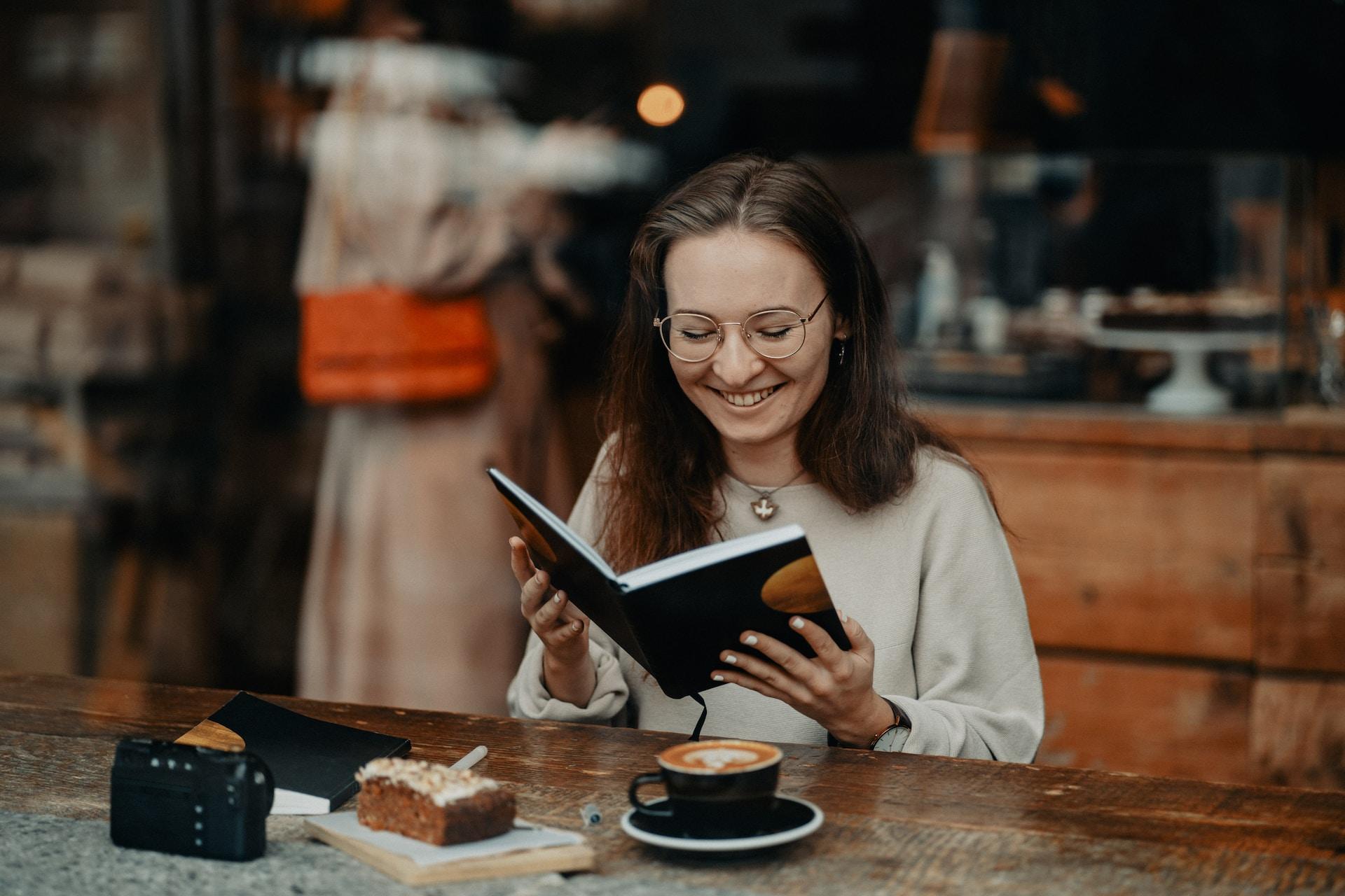A girl sits at a table in a cafe with a coffee and some pastries holding a notebook and smiling.